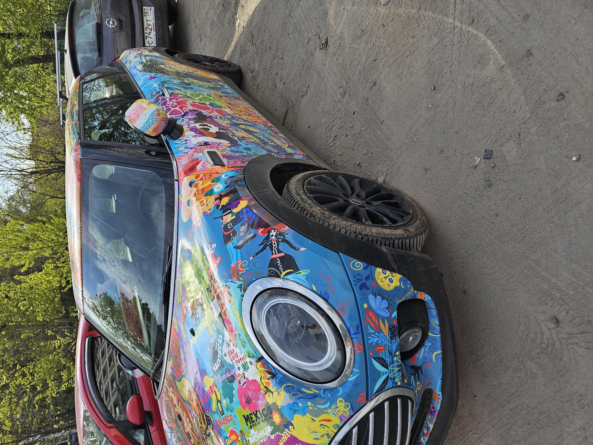 The image shows a brightly decorated Mini Cooper parked on a street. The car is adorned with colorful designs that include images of flowers, people, and text related to various Mexican states and cities, such as Jalisco, Mexico City, Michoacán, and Morelos. The Russian license plate "С742 УТ 197" is visible on a dark car parked behind it. The Mini Cooper has a black wheel and tire. The ground is concrete or asphalt. There are green trees in the background, suggesting the car is in an urban environment in Moscow, Russia.