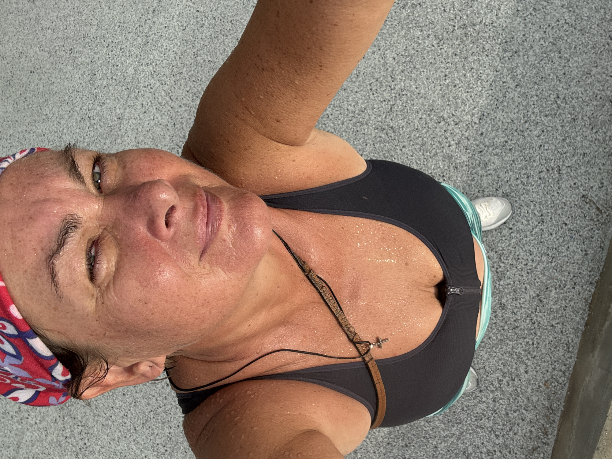 A medium shot shows a sweating woman wearing a headband and sports bra, holding a camera above her head. The woman is fair-skinned with wrinkles. She is wearing a multicolored headband and a gray sports bra. A leather strap with a cross pendant hangs from her neck. A wire wraps around her neck and down the side of her body. The ground is asphalt. One running shoe is visible. The location is not determinable from the image.