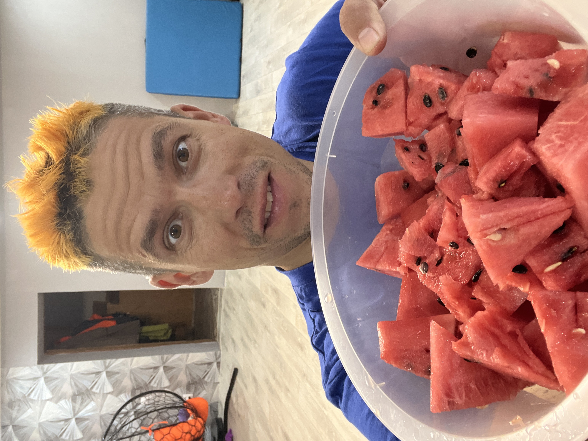 A person with bright orange dyed hair holds a clear plastic bowl filled with sliced watermelon. The person is wearing a blue t-shirt. The bowl of watermelon is positioned in the foreground, partially obscuring the person's face. The person is looking towards the camera with a slightly open mouth and a hint of a smile. Their eyes are brown. The scene appears to be indoors, with a light-colored tiled wall visible behind them. In the background, there is a blue mat on a white surface, suggesting a gym or exercise space. The lighting is bright, indicating it could be daytime. The overall mood seems casual and lighthearted, with the watermelon suggesting a refreshing snack.