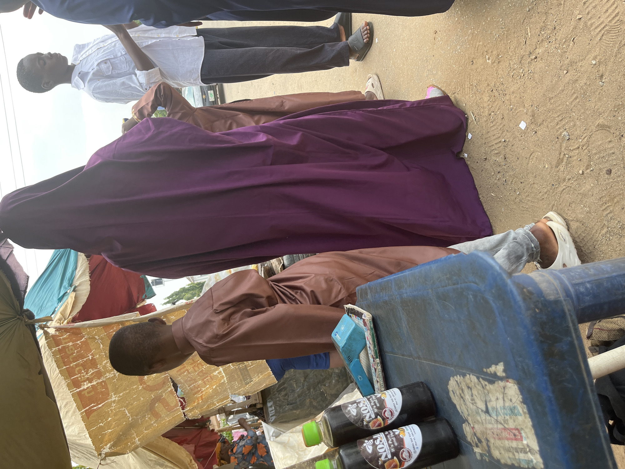 This image, taken in Abuja, Nigeria, shows a scene with several individuals and objects. In the foreground, a person wearing a brown robe is leaning over a blue plastic table. On the table, two dark bottles with green caps are visible, bearing the text "NOSI Organic and Natural It's Pure, Rich and Healthy". There is also a blue electronic device and some paper items on the table.
Behind this individual, a person in a purple garment is also leaning forward. Further back, a young person in a white shirt and dark pants stands, looking upwards. The ground appears to be sandy and dusty. In the background, there are various makeshift structures with colorful fabric coverings, suggesting a market or roadside stall setting. The lighting suggests it is daytime, possibly overcast. The overall impression is one of a bustling outdoor environment, possibly a market, where people are engaged in everyday activities.