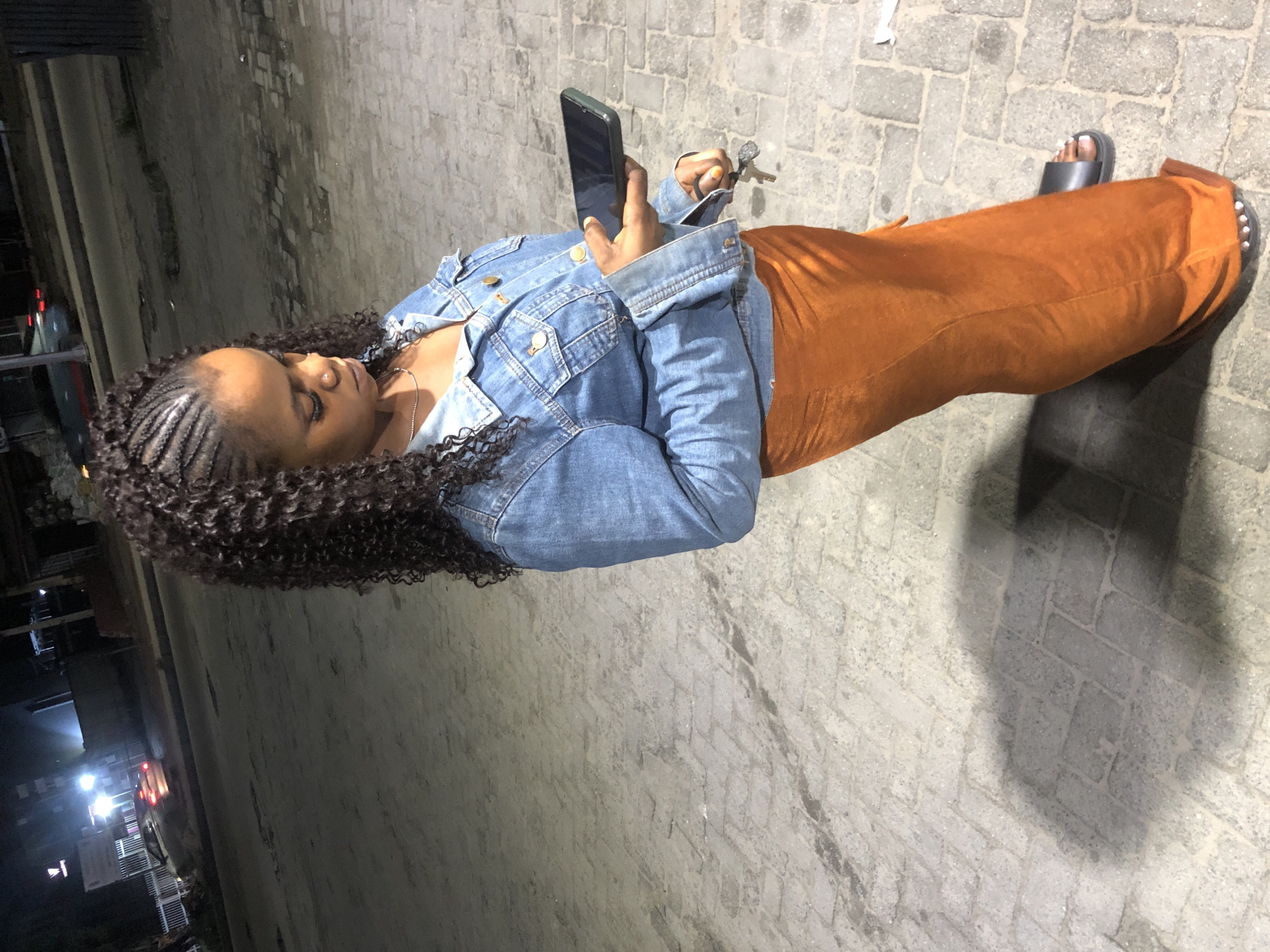 In Ado, Nigeria, at night, a young woman stands on a brick-paved outdoor surface, illuminated by artificial light. She is wearing a denim jacket over a silver necklace and an orange, floor-length velvet skirt. Her hair is styled in braids at the top and has curly extensions. She is looking down at a dark-colored smartphone she holds in her right hand, while her left hand holds a set of keys. The background shows a dimly lit street with a car in the distance and some buildings. A shadow of the woman is cast on the ground.