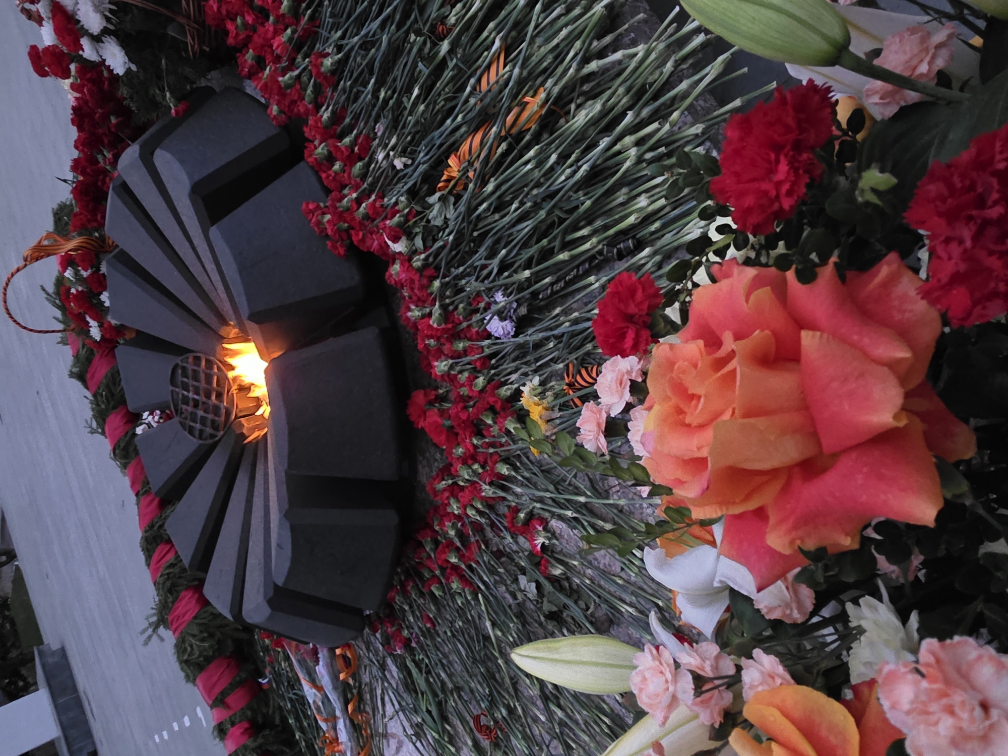 The image shows an eternal flame monument, surrounded by an arrangement of flowers. The monument is constructed from dark gray material and is shaped with angular segments surrounding a central grate through which the flames are visible. The monument is encircled by an arrangement of flowers, primarily red carnations, but also including a large peach-colored rose, white lilies, and other assorted blossoms. A ribbon with black and orange stripes is also visible among the floral arrangements. The setting appears to be a public space, likely in Chaikovsky, Russia. The overall scene conveys a sense of remembrance and respect.