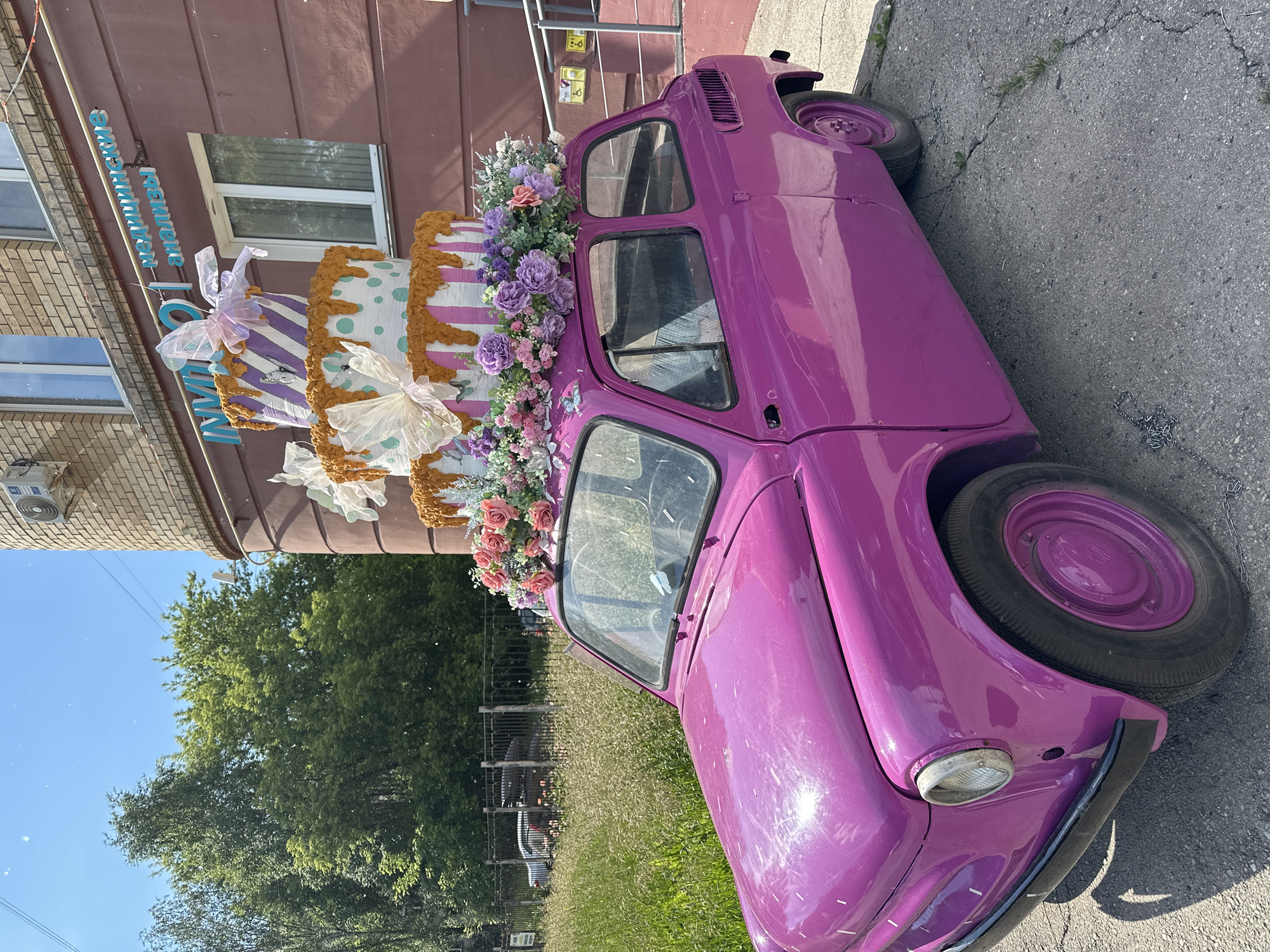 A bright pink, vintage two-door car is parked on asphalt outside a brick building in Perm, Russia. The car is topped with a three-tiered, faux cake decorated with pink and white stripes, teal polka dots, and dripping caramel icing. The cake is adorned with large, sheer pink and white bows, artificial flowers in shades of pink, purple, and white, and small butterfly decorations. Above the cake, a sign on the building reads "INVITRO медицинские анализы" which translates to "INVITRO medical analyses". The building has windows with white frames, and an air conditioning unit is visible on the upper brick wall. To the left of the car, a chain-link fence encloses a grassy area with trees in the background, under a clear blue sky. The car's headlights and chrome trim are visible, as well as its round purple hubcaps.