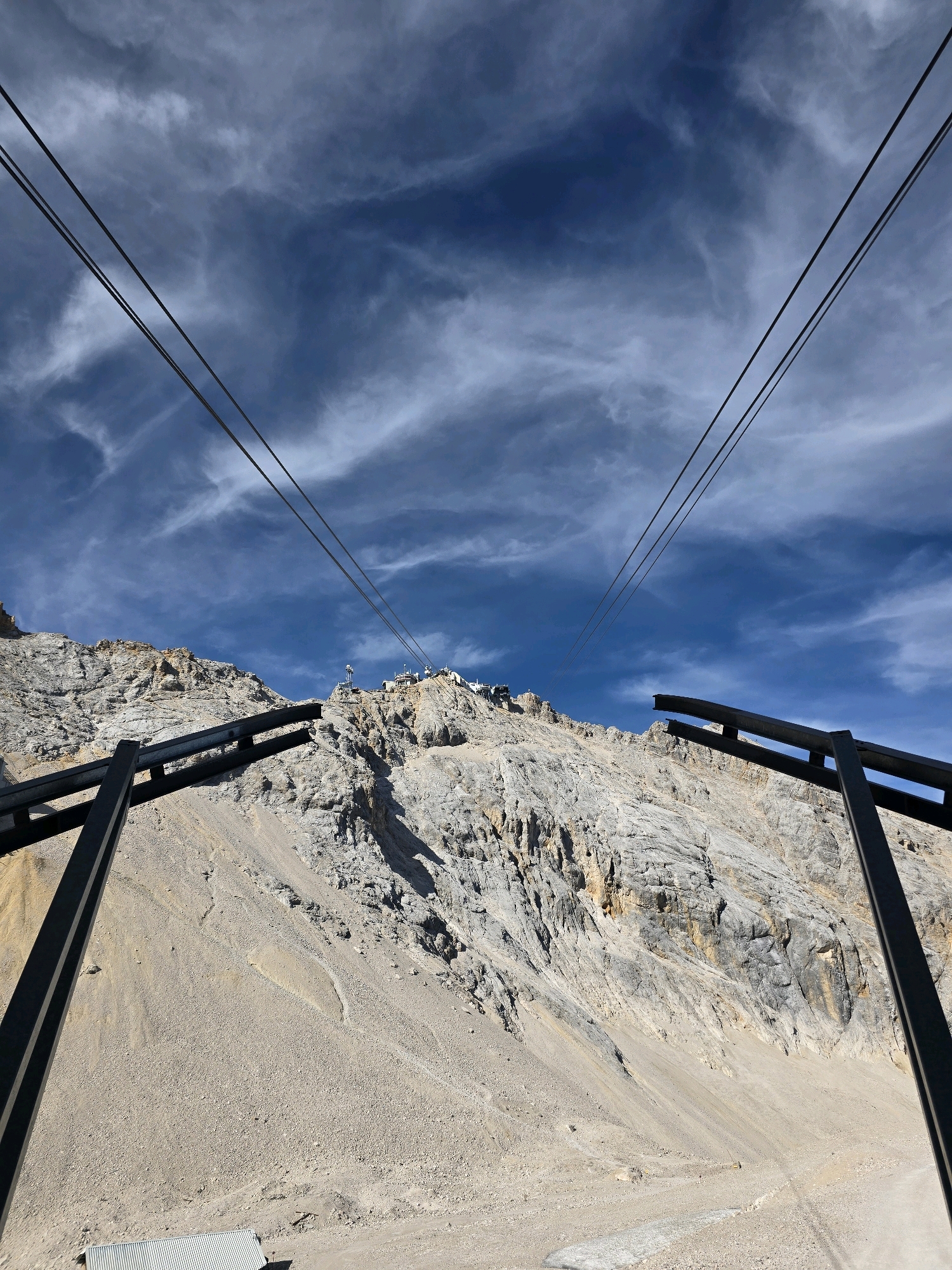 This is a wide-angle, low-angle shot looking up at a steep, rocky mountain face with sparse vegetation and a bright blue sky filled with wispy clouds. In the foreground, two large, black metal structures frame the view of the mountain. Multiple parallel cable car wires stretch across the sky towards the mountain summit, where a complex of white buildings is visible. The ground leading up to the mountain is covered in loose gravel and scree. The scene appears to be the Zugspitze, Germany's highest peak, located near Garmisch-Partenkirchen. There are no people visible in the image.