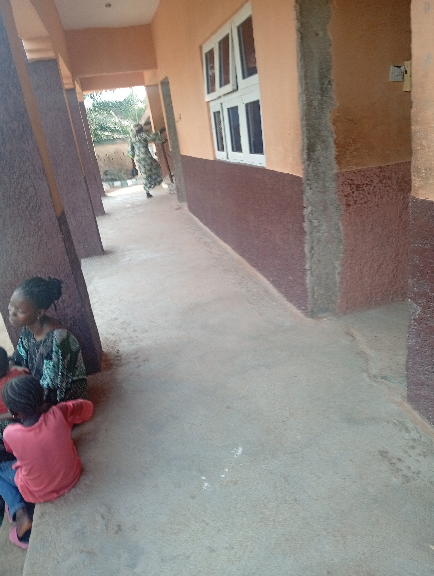 The image shows a group of people outside a building in Jalingo, Nigeria. In the foreground, two young children are sitting on the concrete ground. One child is wearing a pink t-shirt and blue jeans, and the other is partially visible, wearing blue jeans and pink slippers. A woman wearing a patterned dress is sitting next to them, looking to the side. In the background, another woman in a patterned dress is walking away from the camera down a covered walkway. The walkway has a light-colored concrete floor and is flanked by columns with a textured, reddish-brown finish. The building has peach-colored walls with a band of darker reddish-brown at the bottom. A row of white-framed windows is visible on the building's exterior. The scene appears to be outdoors during the daytime, with some vegetation visible in the distance. The overall impression is of a casual gathering in a public space. There is no visible text in the image.