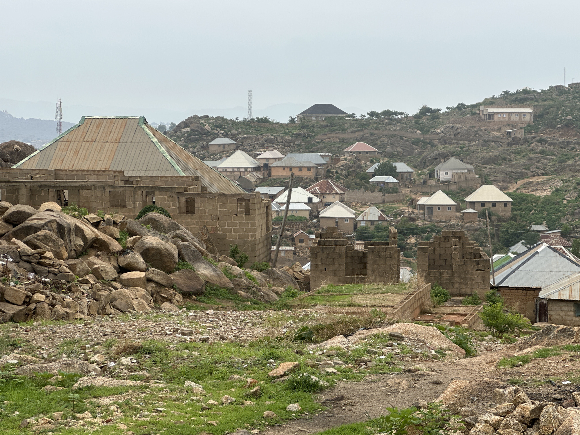 The image shows a neighborhood in Jos, Nigeria. In the foreground is a rocky, uneven terrain with sparse green vegetation. Several unfinished or abandoned concrete block structures are visible, mixed with piles of rocks. In the background, numerous houses with corrugated metal roofs of various colors are scattered across a rocky hillside. Further back, communication towers are faintly visible against a hazy sky. The overall scene suggests an underdeveloped or impoverished residential area with ongoing construction amidst a rugged landscape.