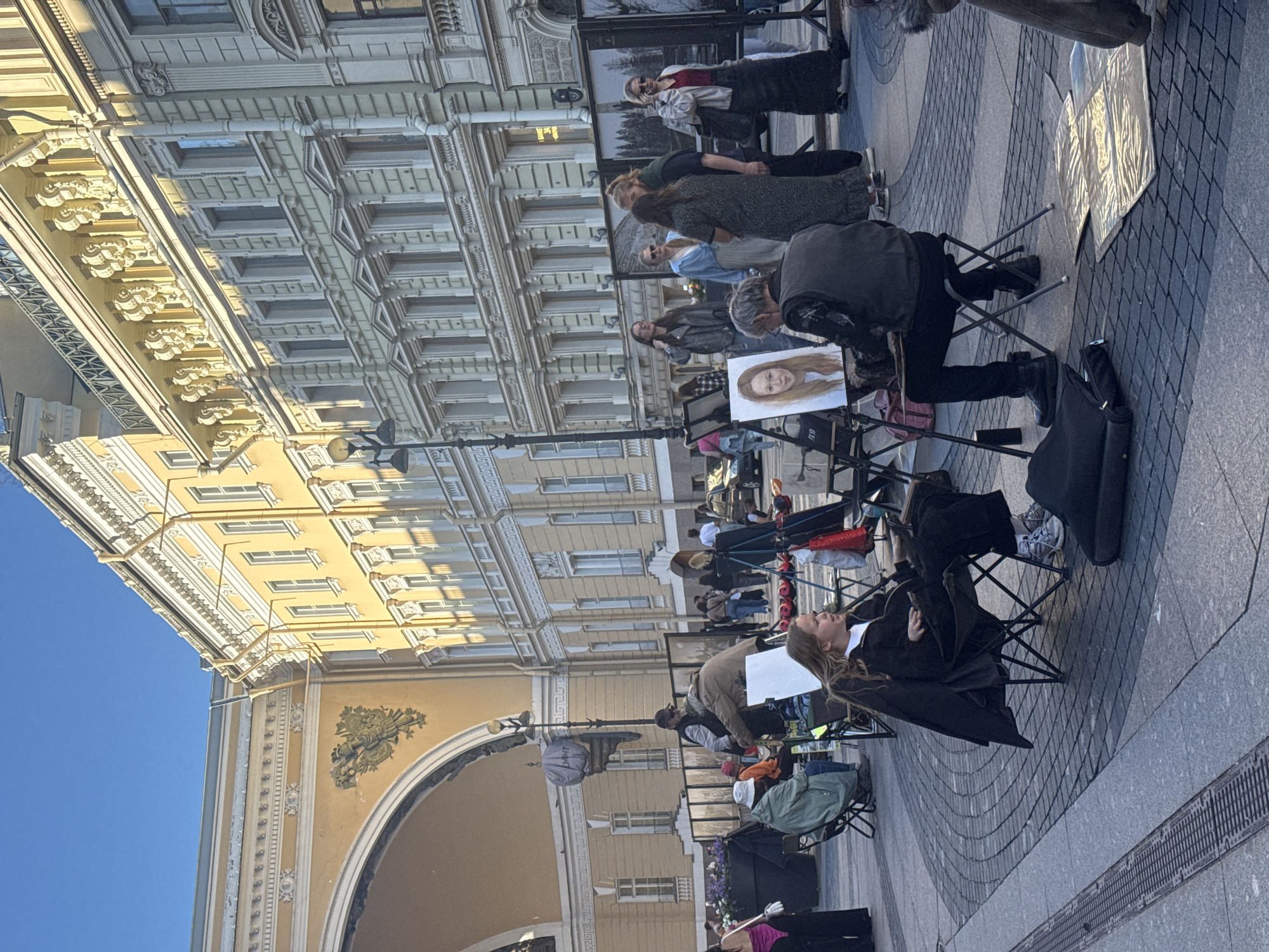 This image depicts an outdoor art scene in Palace Square, Saint Petersburg, Russia, with the General Staff Building in the background. Several artists are set up, displaying their work and offering portrait sittings.
In the foreground, a young woman with blonde hair is seated in a black folding chair, looking to her right. A male artist, wearing a dark vest and pants, is seated on a stool facing her and appears to be in the process of painting her portrait on an easel. A finished portrait of a young woman is visible on another easel.
Behind them, more artists and onlookers are present. Several large framed photographs or paintings are displayed on stands. People are browsing the artwork and interacting with each other. The paving stones of the square are visible underfoot, and architectural details of the surrounding buildings, including ornate facades and arched structures, are prominent. The scene is bathed in natural sunlight, suggesting it is daytime.