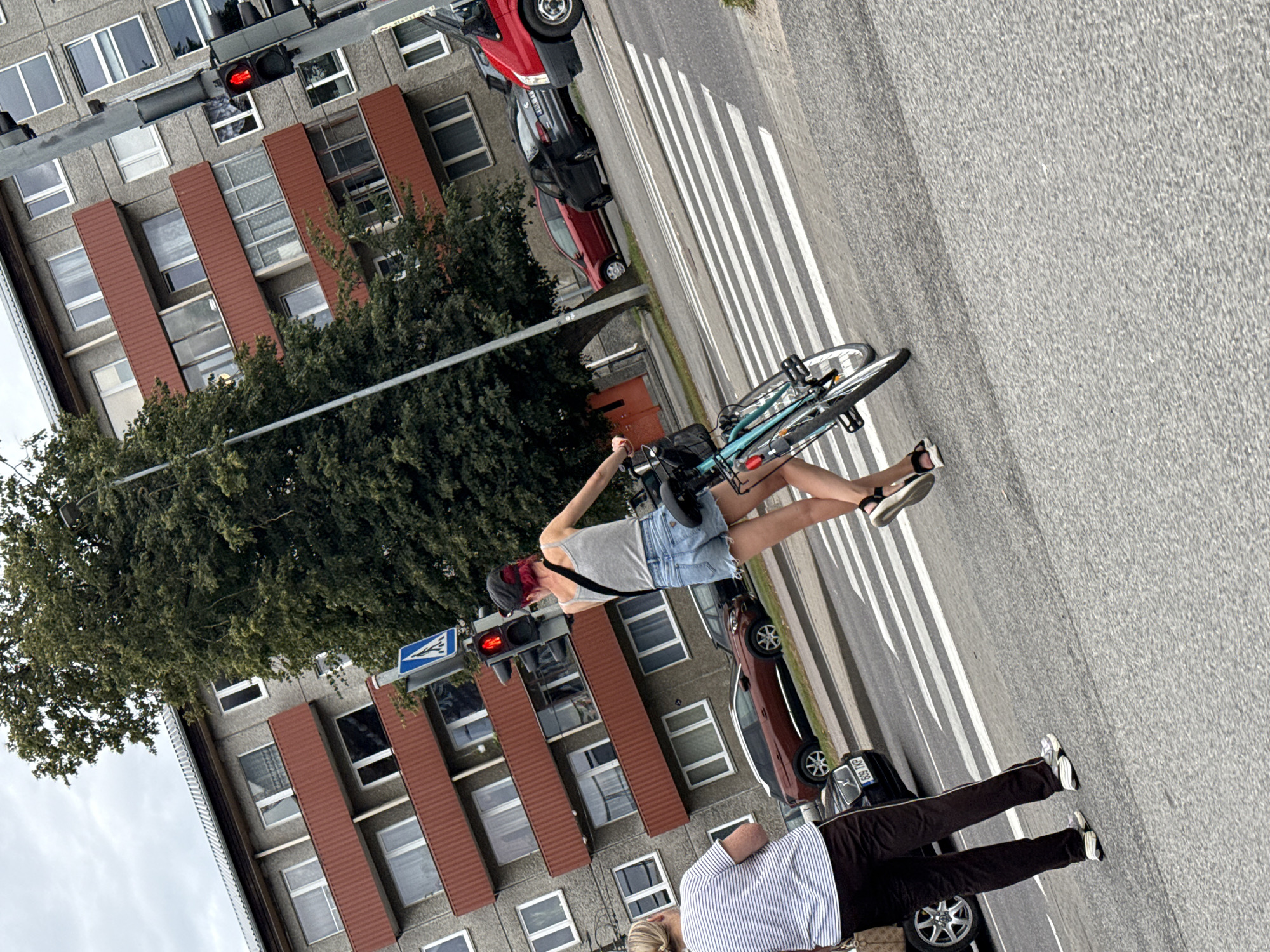 Two individuals are at a crosswalk in an urban environment. The person on the left, facing away from the camera, wears a striped t-shirt and dark pants. They are standing on the asphalt. The person on the right, also facing away from the camera, is pushing a bicycle. They are wearing a grey tank top and denim shorts. The bicycle is light blue and has a basket on the front. Both individuals are positioned on the white stripes of the crosswalk. In the background, a multi-story building with red-trimmed balconies and numerous windows is visible. Traffic lights are present, with one showing a red signal. Several vehicles, including a red van and a dark-colored car, are on the road. A large tree with dense foliage stands to the right of the building. The sky is overcast.