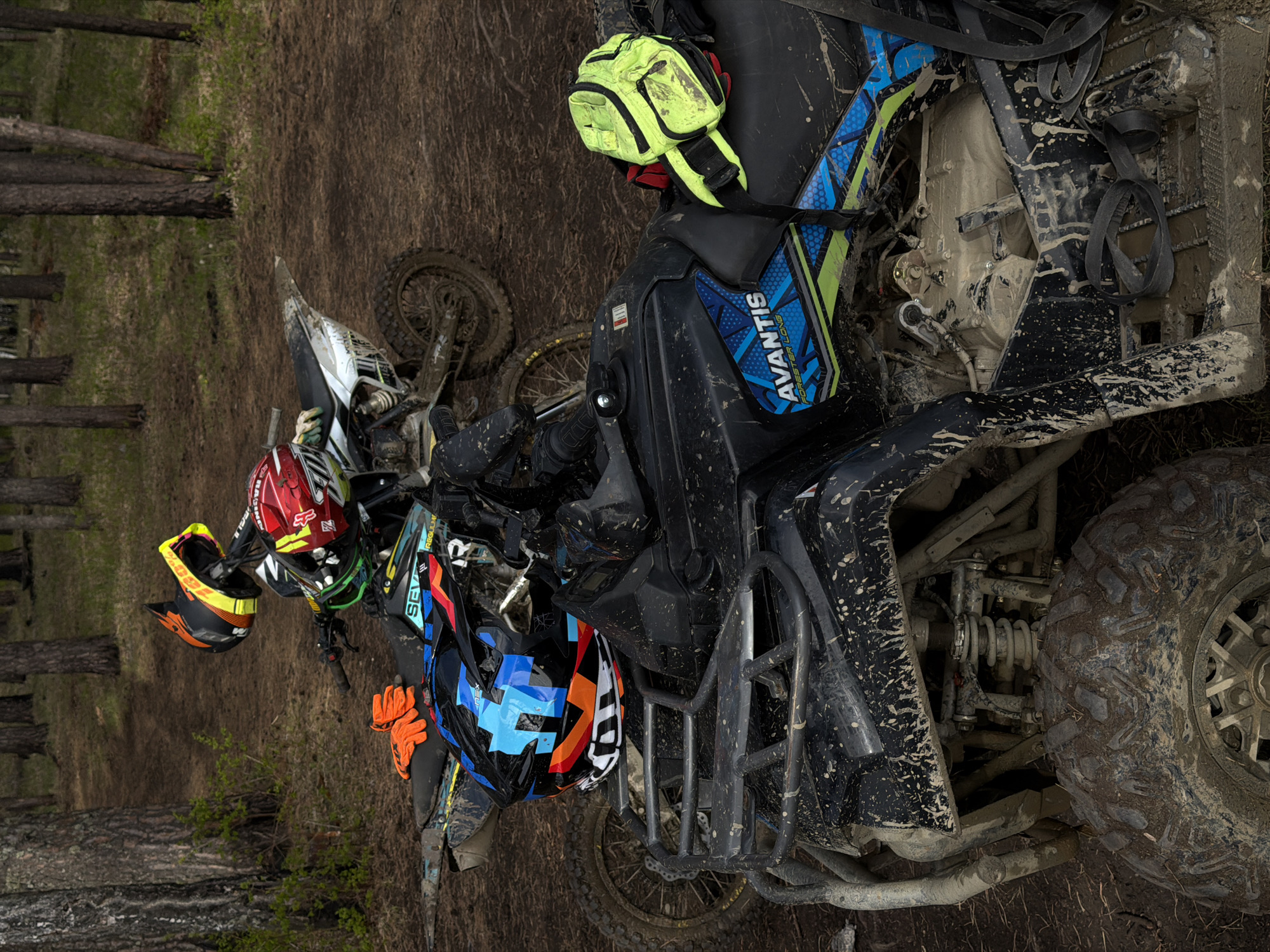 The image shows an ATV and a dirt bike in a muddy, wooded area, possibly near Манский, Russia. Both vehicles are covered in mud. Several helmets and gloves are also visible. The ATV is branded "AVANTIS." The background consists of trees and muddy ground.