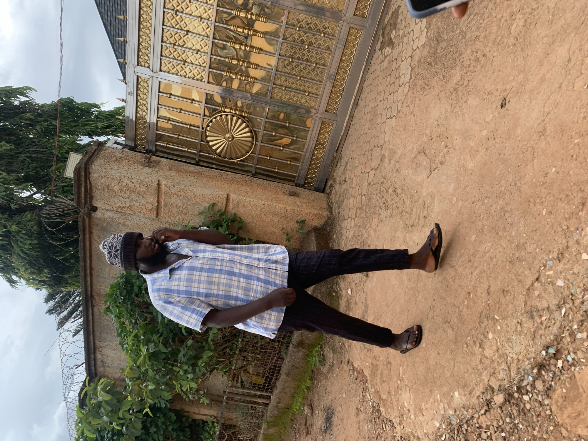 A man walks on a reddish-brown dirt path under an overcast sky in Jos, Nigeria. He is of African descent, has a beard, and wears a light blue and white plaid short-sleeved shirt, dark pinstriped trousers, and black flip-flops. On his head, he wears a white, grey, and dark brown patterned knit beanie. He holds a dark-colored smartphone to his right ear, seemingly engaged in a call, as he moves from right to left in the frame. To his left, a textured brownish-tan wall topped with razor wire is partially obscured by dense green foliage. To his right, a large, ornate golden and dark grey metal gate features intricate patterns, including a central circular sunburst design. Above the gate, a dark-colored roof and tall green trees are visible, with a light fixture mounted on the wall post next to the gate. The ground transitions from unpaved dirt to a section of patterned paving stones closer to the gate. A hand holding a smartphone is partially visible in the bottom right corner.