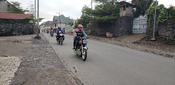 The image shows a street scene in Keshero, Democratic Republic of the Congo. There are three motorcyclists traveling on an asphalt road, which is bordered on both sides by a rock wall. The road is in poor condition, with areas of uneven pavement and loose gravel. The motorcyclists are wearing helmets and are all traveling in the same direction. There are a few small buildings and trees on the sides of the road. The motorcyclists are traveling on the right side of the road. One of the motorcyclists is wearing a green shirt with the word “STRATEGIC” on it. There is a metal gate in the background, with the words “XXIX” on it. The scene is a common sight in Keshero, where motorcycles are a popular mode of transportation.