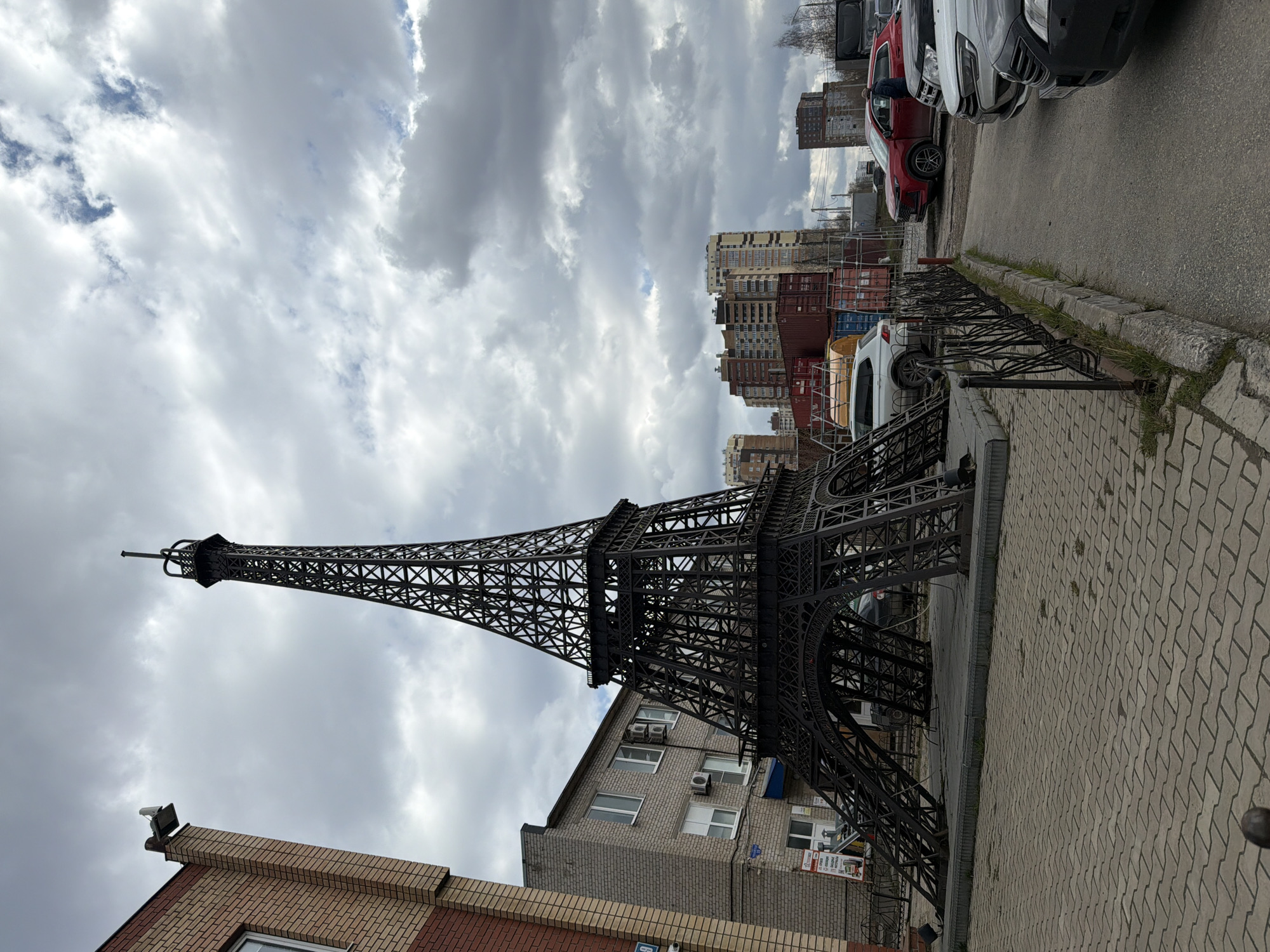 The image displays a large, dark metal replica of the Eiffel Tower situated in an urban environment. The tower stands on a paved sidewalk, constructed from interlocking concrete pavers, alongside a curb and an asphalt road.
In the foreground, to the left, is a building with a mixed red and lighter-toned brick facade. Directly behind the base of the Eiffel Tower replica is another multi-story building, constructed from gray brick, featuring numerous windows and several exterior air conditioning units. Further in the background, a cluster of taller residential or commercial buildings can be seen, adding to the urban landscape.
Along the side of the asphalt road, several cars are parked, including a prominent red car, a white car, and other vehicles partially visible. Fencing and what appear to be large industrial containers or storage units are present in the mid-distance on the right.
The sky is heavily overcast with a mass of grey and white clouds, indicating a cloudy daytime. No individuals are visible in the scene. A sign with unreadable Cyrillic text is visible on the gray brick building directly behind the tower replica. The overall setting is characteristic of a city street, with the Eiffel Tower replica serving as a decorative landmark.