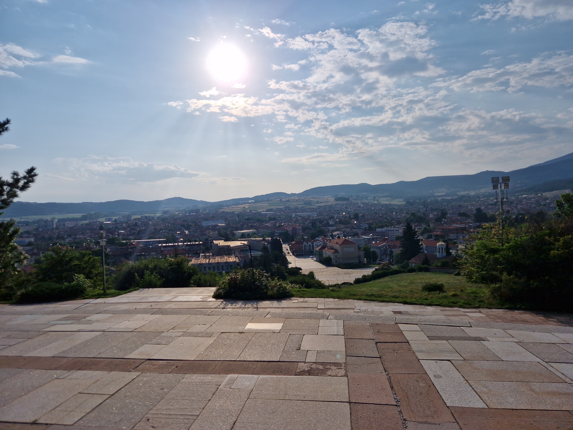 The image is an outdoor, high-angle shot overlooking the city of Panagyurishte, Bulgaria, on a clear, sunny day. In the foreground, a wide, paved area made of large, rectangular stone tiles in various shades of grey and brown leads the viewer's eye towards the city. Some tiles appear worn or damaged. A dark green, bushy plant is situated to the left of the tiled area, and a sparse conifer tree branch extends from the upper left corner of the frame.
The middle ground features a densely populated urban landscape with numerous buildings characterized by red-tiled roofs and some lighter-colored facades. A large, gold-domed building stands out among the structures. Green trees and vegetation are scattered throughout the city, and a wider, paved public space is visible in the mid-distance. Two tall, metal light poles with multiple fixtures are positioned on a grassy incline to the right of the city center.
The background consists of rolling, tree-covered hills that frame the city. The sky is a bright blue with scattered, fluffy white clouds. The sun, a bright, intense orb, is visible in the upper left quadrant of the sky, casting rays of light that penetrate the clouds and illuminate the scene. No people are visible in the image. The overall impression is one of a peaceful, sun-drenched afternoon in a Bulgarian town.
