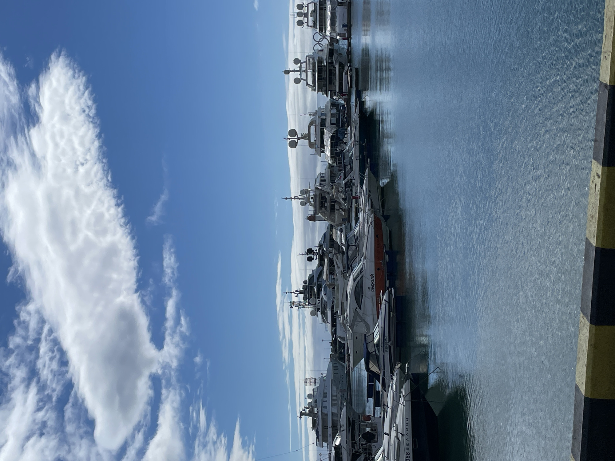 The image shows a marina in Sochi, Russia. Several yachts of varying sizes are docked along a pier. The sky is blue with scattered white clouds. The water is calm and reflects the sky and the yachts. A section of the pier with black and yellow markings is visible on the right side of the image.