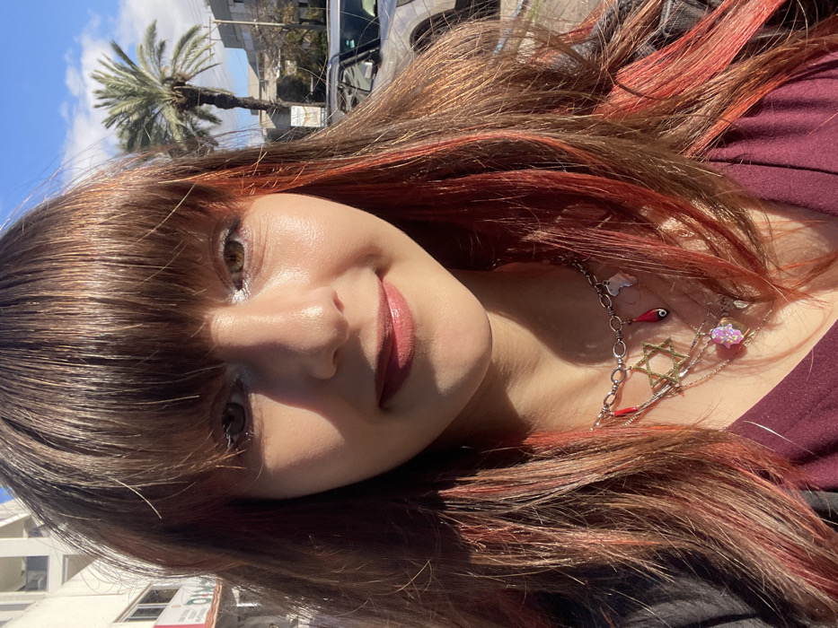 A young female subject is depicted in a head-and-shoulder portrait, positioned centrally and looking directly at the camera with a subtle smile. Her long, dark brown hair features prominent reddish-orange highlights, especially on the right side, and includes bangs across her forehead. Her eyes are light brown, with white eyeliner applied to the lower lash line. She wears light pink lipstick or gloss. Multiple layered necklaces adorn her neck; a silver-colored chain displays several charms including a golden Star of David, two red teardrop-shaped pendants (one with an eye motif), a small translucent charm shaped like the continent of Africa, and a pink iridescent Hamsa hand. She is wearing a maroon-colored top with a dark outer garment visible on her left shoulder. The background is softly blurred, revealing an urban setting under a clear blue sky with scattered white clouds. A tall palm tree stands on the right side of the frame, with buildings featuring balconies visible on the far left. A light-colored vehicle is partially discernible further in the background. The scene is an outdoor street environment in Raanana, Israel, under natural daylight.