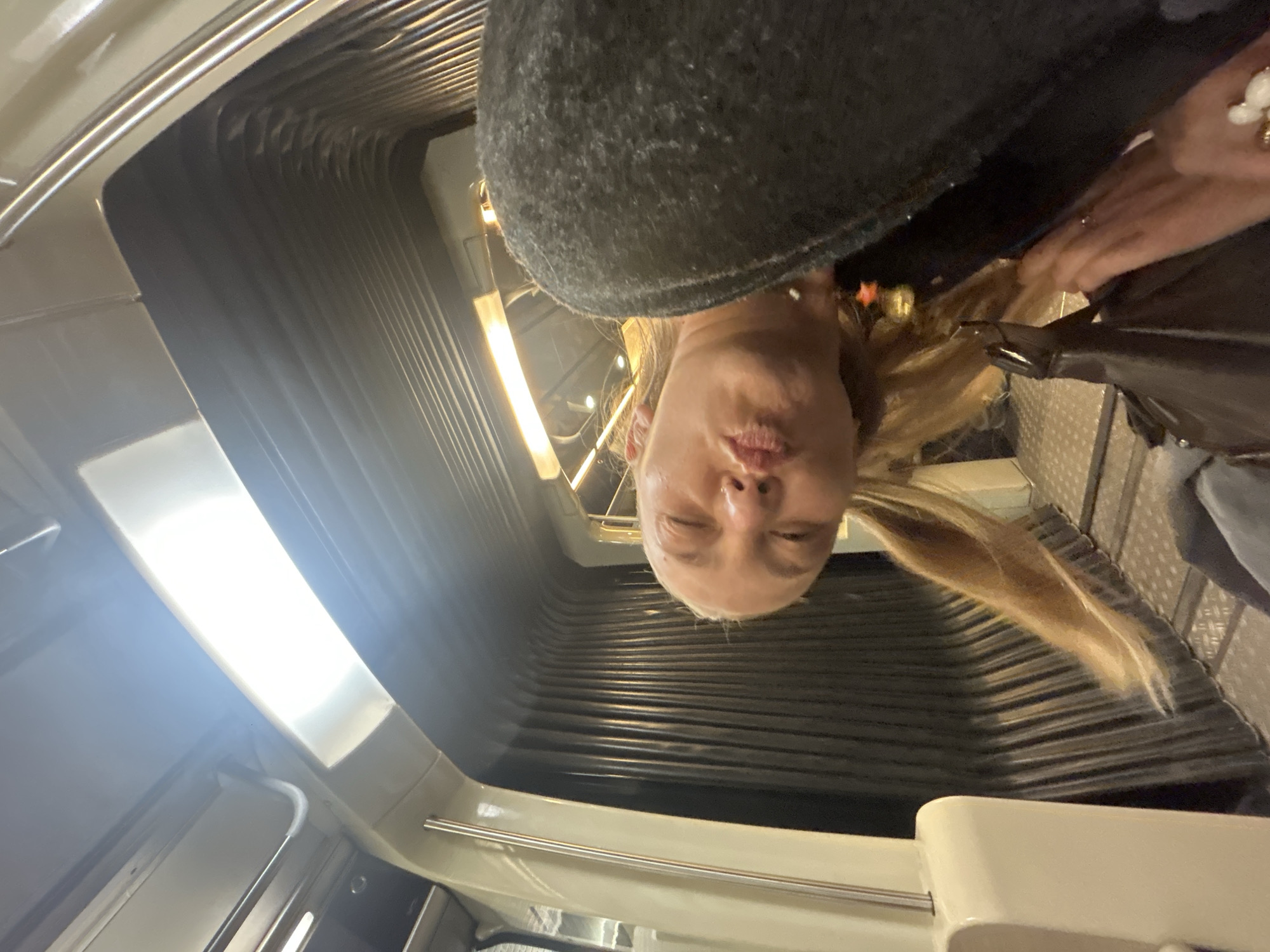A woman is pictured inside a train car, seemingly on the Paris Metro, based on the interior of the train car. The woman has blonde hair, is wearing a gray hat, and is looking directly at the camera with her lips pursed. The train car has gray ribbed walls and a window with a light above it.