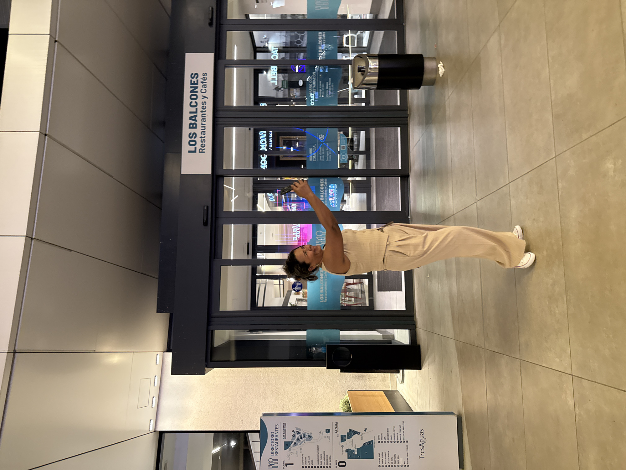 A woman stands on a tiled floor in front of a glass-fronted building with a sign that reads "LOS BALCONES Restaurantes y Cafés." She is wearing a tan, sleeveless jumpsuit and white sneakers. The woman is holding a cell phone in her right hand and appears to be taking a photo or video. The scene is indoors, likely in a mall or shopping center, indicated by the directory map visible at the bottom of the image which lists "TresAguas" and specific restaurant names. The directory map also shows floor plans for "PLANTA 1" and "PLANTA 0" and provides opening hours. The lighting suggests it is daytime. The overall mood appears casual and relaxed.