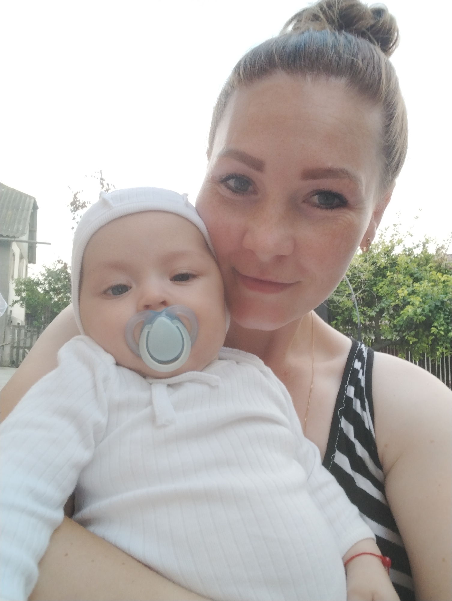 In Fălești, Moldova, a medium shot captures a woman holding a baby. The woman, with her blonde hair pulled up in a bun and visible freckles on her face, looks towards the camera with a slight smile. She is wearing a black and white striped tank top and a thin gold necklace. The baby, swaddled in a white ribbed onesie and a matching white hat, is being held close to the woman. The baby has a light blue pacifier in its mouth. Behind them, the blurred background shows a residential building with a grey roof and some green foliage. The lighting suggests it is daytime, possibly outdoors.