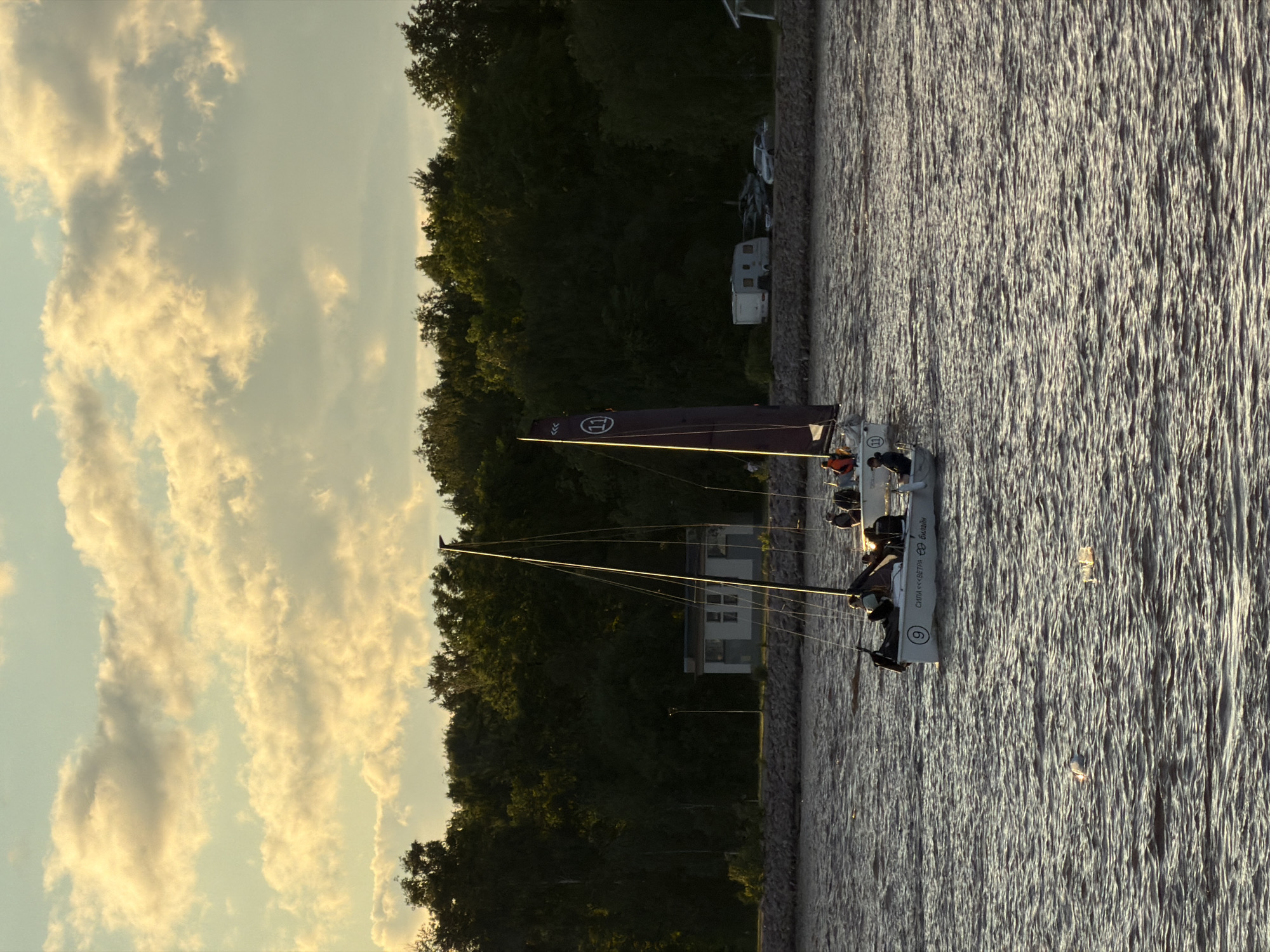 This is a photograph of a sailing race on a body of water, likely the Gulf of Finland, near Saint Petersburg, Russia. The image captures several sailboats with crews navigating the water. The sky is filled with dramatic, golden-lit clouds, suggesting either sunrise or sunset.
In the foreground, the choppy water reflects the light. Two sailboats are prominently featured. The one closest to the viewer has a white hull with the number "9" in a black circle and Cyrillic text that appears to read "СИЛА <<< ВЕТРА >>> БИЛЬБАЙН." Several individuals are visible on this boat, dressed in life vests.
Slightly further back and to the right, another sailboat is visible, its dark purple sail displaying the number "11" in a white circle. This boat also has a crew onboard.
In the background, a line of dense green trees forms the shoreline. Behind the trees, a small, light-colored building with windows and a dark roof is visible. Further to the right along the shore, a white recreational vehicle or caravan is parked, along with several cars. A stone embankment or wall runs along the water's edge. The overall atmosphere is one of a calm, yet active, maritime scene set against a beautiful sky.