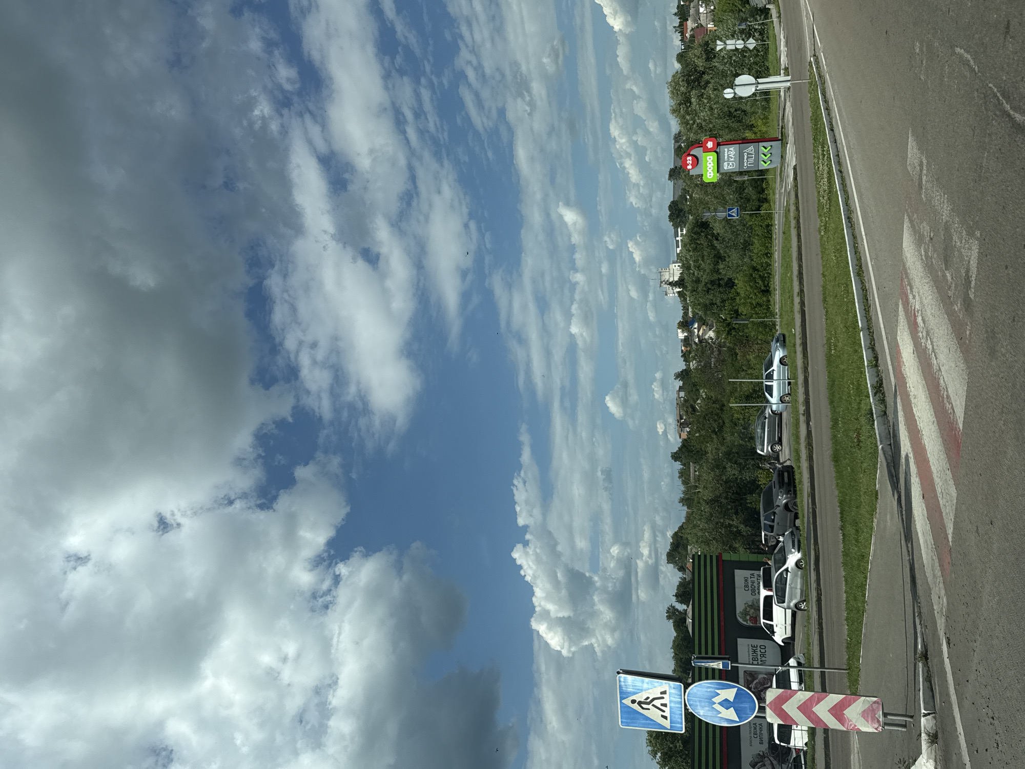 The image shows a roadside scene in Skvyra, Ukraine, on a sunny day with scattered clouds. A paved road runs alongside a grassy area with trees. Several cars are parked along the side of the road, and a pedestrian crossing is visible. There are also various signs. One sign displays "8-23" and logos for "ФОРА", "КАВА", and "ПІЦА", indicating a store with these offerings and operating hours. Another sign shows directional arrows, and a blue sign with a pedestrian symbol indicates a crosswalk. A red and white chevron-patterned sign warns of a directional change. The overall impression is that of a typical day in a Ukrainian town.
