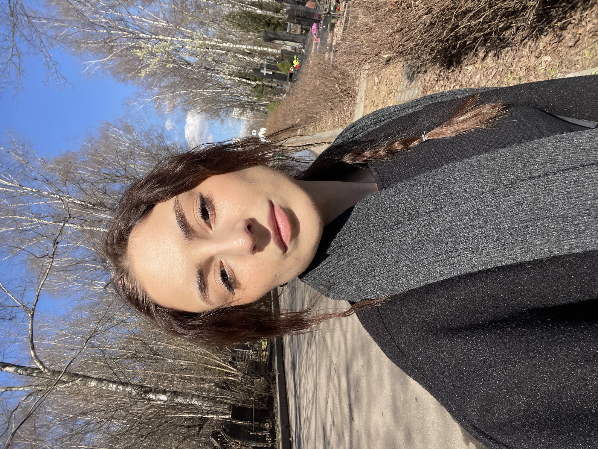 A vertical outdoor head and shoulders portrait shows a young woman with brown hair wearing a black top and gray scarf. She is angled to the right, facing forward. Her hair is styled with a braid. The scene is set in a graveyard in Moscow, Russia. Bare trees line the background, and tombstones are visible in the distance. The sky is blue. A path is seen along the right side of the image.