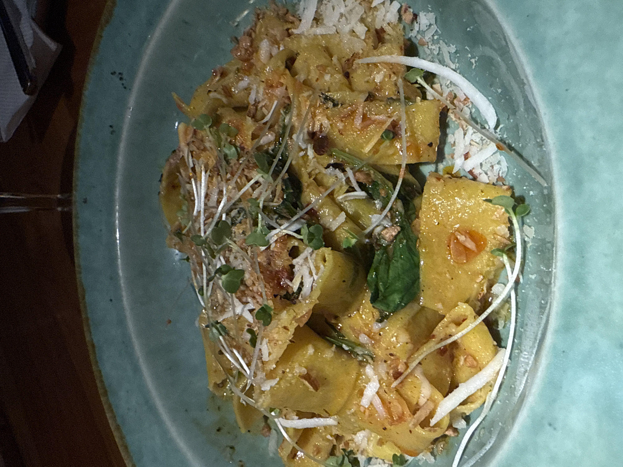 The image shows a plate of pasta on a light blue-green ceramic plate. The pasta is yellow and appears to be a larger, square cut, topped with shredded cheese or parmesan, fried garlic, and microgreens. A portion of the plate is visible, showing a glossy finish. In the upper left corner, there is a glimpse of a dark brown table and possibly a napkin and cutlery. The overall scene suggests a restaurant setting. Since the location is Panaji, India, it is likely a restaurant in that city.