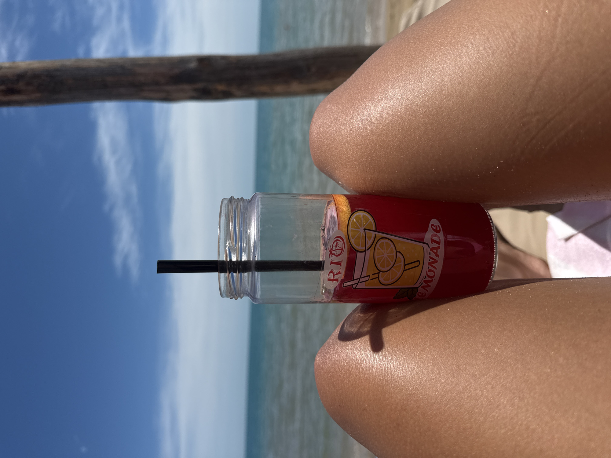 An extreme close-up shot focuses on a person's tanned legs on a beach, with a jar of red lemonade featuring a cartoon image of lemonade and text reading "RIO" and "LEMONADE" positioned between them. The jar is held by a black straw. In the background, the turquoise sea meets a hazy sky. A weathered wooden post is visible on the right side of the frame. The image appears to be taken from a low angle, looking up at the subject and the surrounding environment.