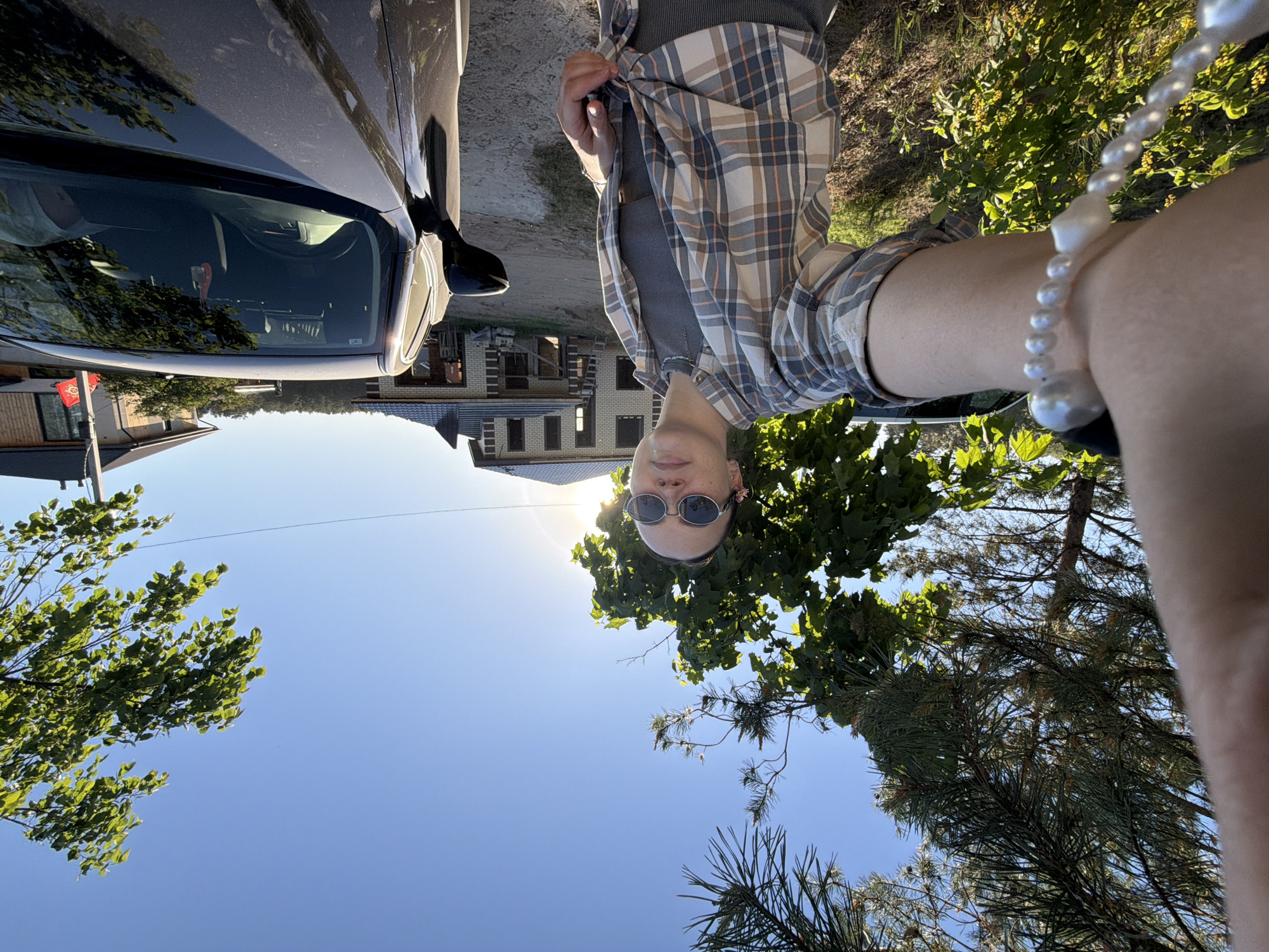 A selfie-style photograph, rotated 180 degrees, shows a person standing next to a car. The person is wearing sunglasses, a gray top, and a plaid shirt tied at the waist. A white pearl bracelet is visible on the person's wrist. The car, partially visible on the left, appears to be a dark color. Behind the person and the car, there is a building under construction. Trees and a blue sky are visible in the background. Based on this information, this image was captured in Chaikovsky, Russia.