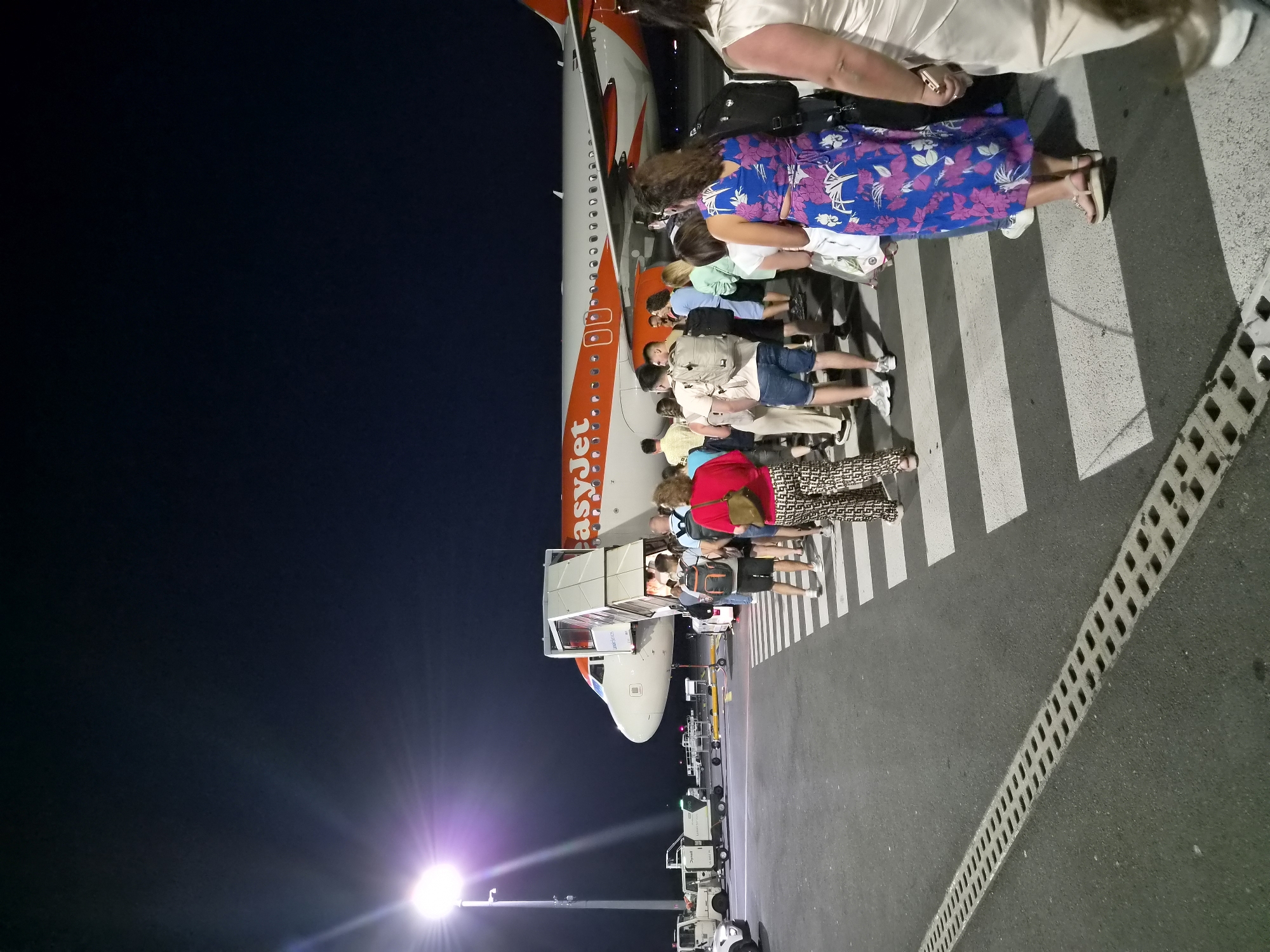 An EasyJet airplane is parked on the tarmac at night. A line of passengers, wearing casual clothing and carrying luggage, are walking across a zebra crossing towards the aircraft's stairs. In the background, several airport service vehicles are visible. A bright floodlight illuminates the scene, casting long shadows. The location is identified as Niederanven, Luxembourg.