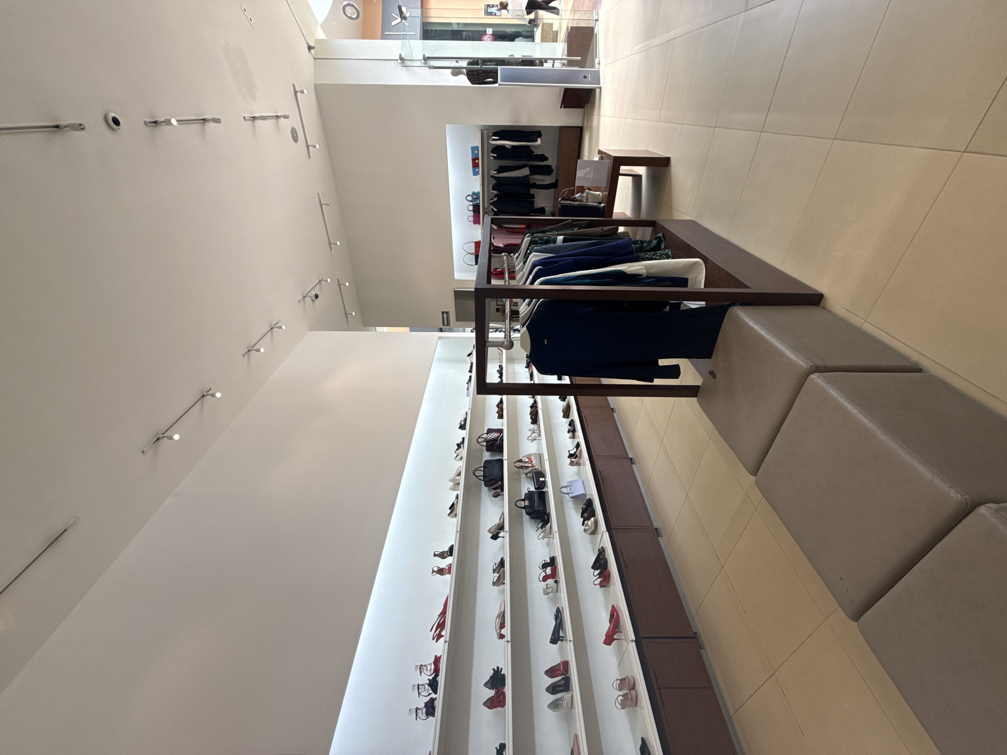 The image shows the interior of a retail store. The store appears to sell clothing, shoes, and accessories. Clothing is displayed on racks near the center of the image, while shoes are displayed on shelving units along the wall to the left. There is a seating area along the right side of the image, consisting of a row of upholstered blocks. The location is likely San Francisco Ocotlán, Mexico. The store appears well-lit with track lighting on the ceiling. No people are clearly visible in the foreground.