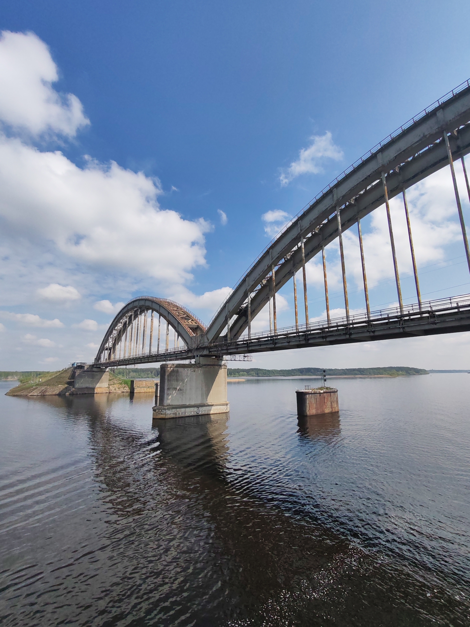 A long, weathered steel bridge spans a wide body of water under a bright blue sky with scattered cumulus clouds. The bridge structure includes two arched sections supported by concrete pillars. The water is dark and rippled, reflecting the sky and the bridge. A concrete structure with a small signal post sits in the water near the bridge. The distant shoreline is visible with a line of trees. This scene is located in Gladyshevo, Russia.