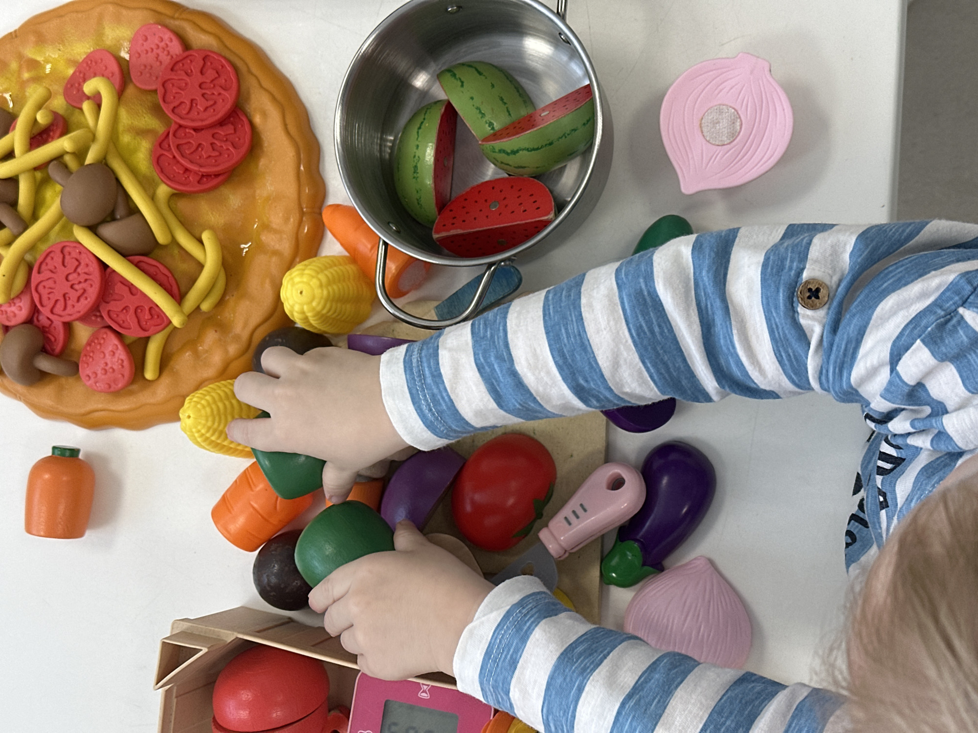 A child wearing a blue and white striped shirt is playing with toy food on a white surface. The child's hands are interacting with various toy fruits and vegetables, including corn, carrots, green avocados, red tomatoes, and purple eggplants. Some of the toy food is arranged in a metal pot, while other pieces are near a toy pizza. There is also a toy timer. The location, Votkinsk, Russia, cannot be determined from the visual information provided.