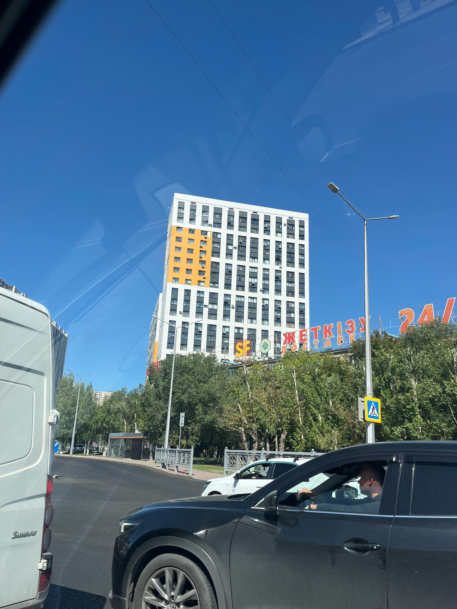 An exterior medium shot shows a bright, sunny day in Astana, Kazakhstan, with a clear blue sky. In the mid-ground, a tall, modern white building with orange accents dominates the scene. The building has many windows and air conditioning units. To the right of the building, a large red sign with white lettering reads "СЕТКІЗЫ 24/". A lamppost with two lights stands near the sign, and a pedestrian crossing sign is visible at its base. Trees with green foliage line the street in front of the buildings. In the foreground, a dark-colored car is partially visible, with a driver inside. A white van is on the left side of the frame. The street appears to be busy with traffic. Reflections of clouds and possibly power lines are visible on the windshield of the car in the foreground.