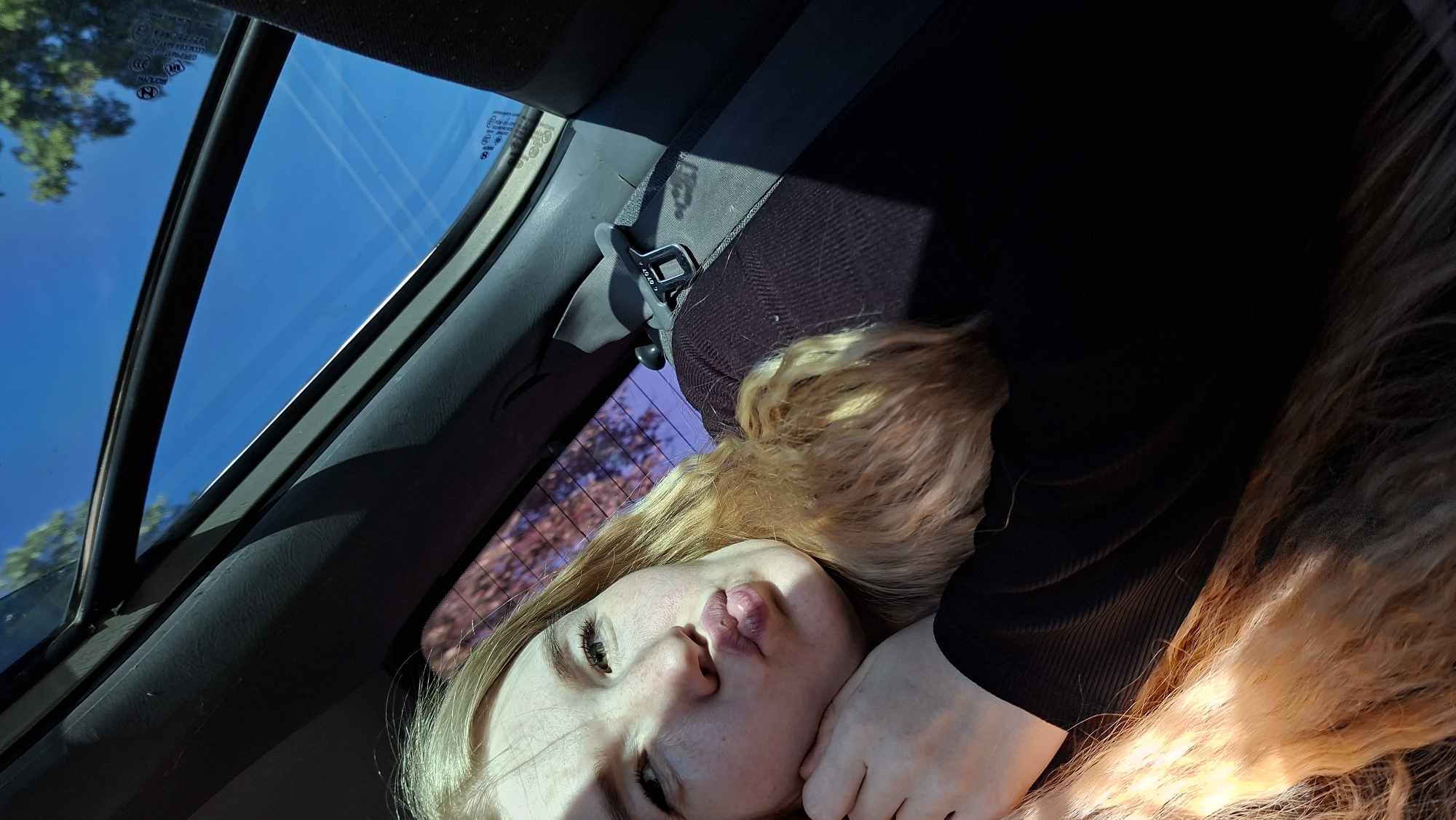 A young woman is lying in a car in Chaikovsky, Russia. The car has a sunroof, and the blue sky is visible through it. A seatbelt is visible across the woman's body. She has long blond hair, is wearing a black shirt, and is pouting. The trees visible through the back window suggest an outdoor environment.