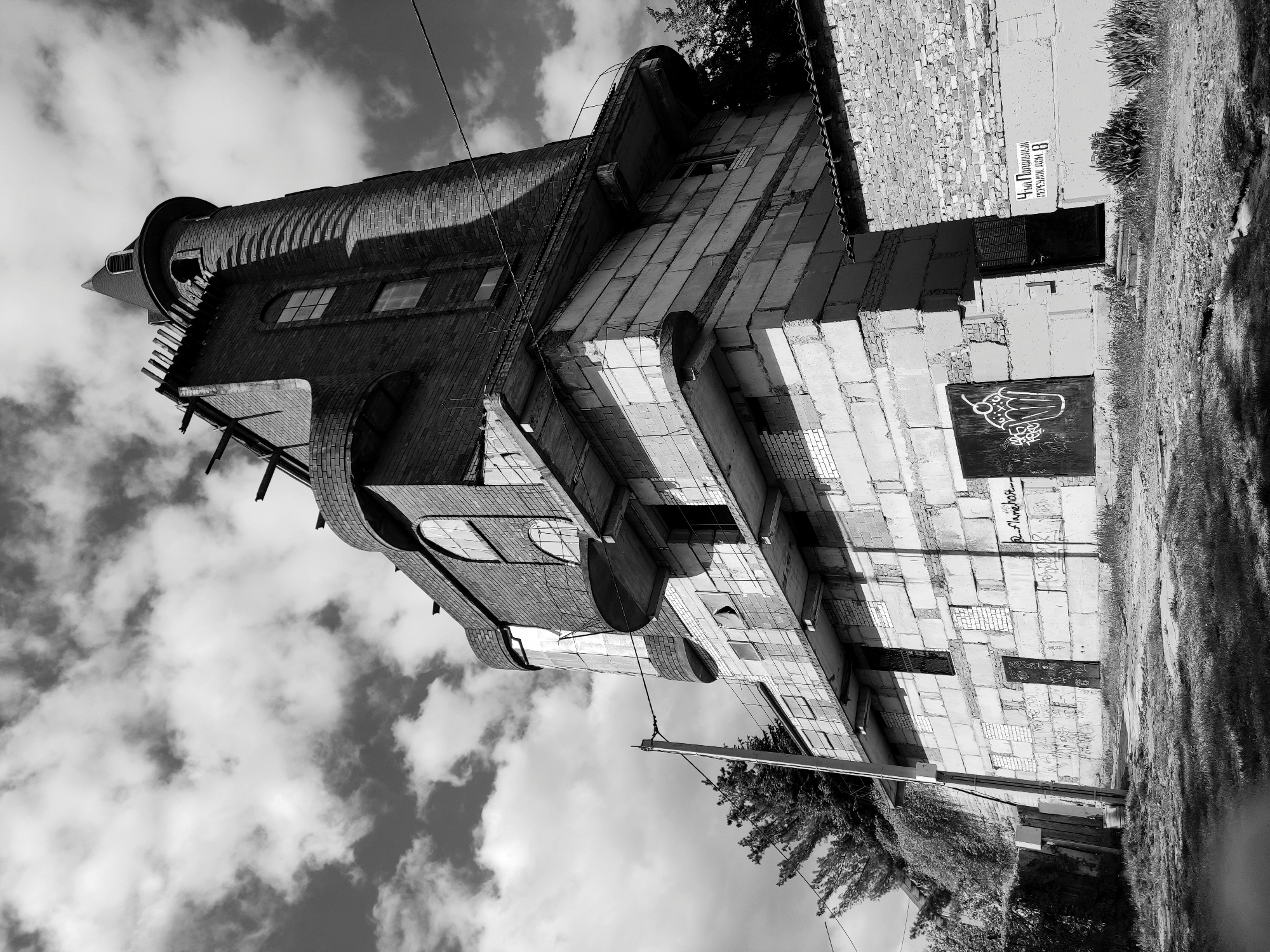 The image shows a black and white, upward-angled shot of a building in Hrodna, Belarus. The building has a tall, cylindrical brick tower attached to a lower, blockier structure. The tower has arched windows and a conical roof. The lower structure is made of large concrete blocks. There is graffiti on one of the walls. A sign in Russian is visible near a door. There are trees and grass visible in the foreground. The sky is filled with clouds. There are power lines visible.