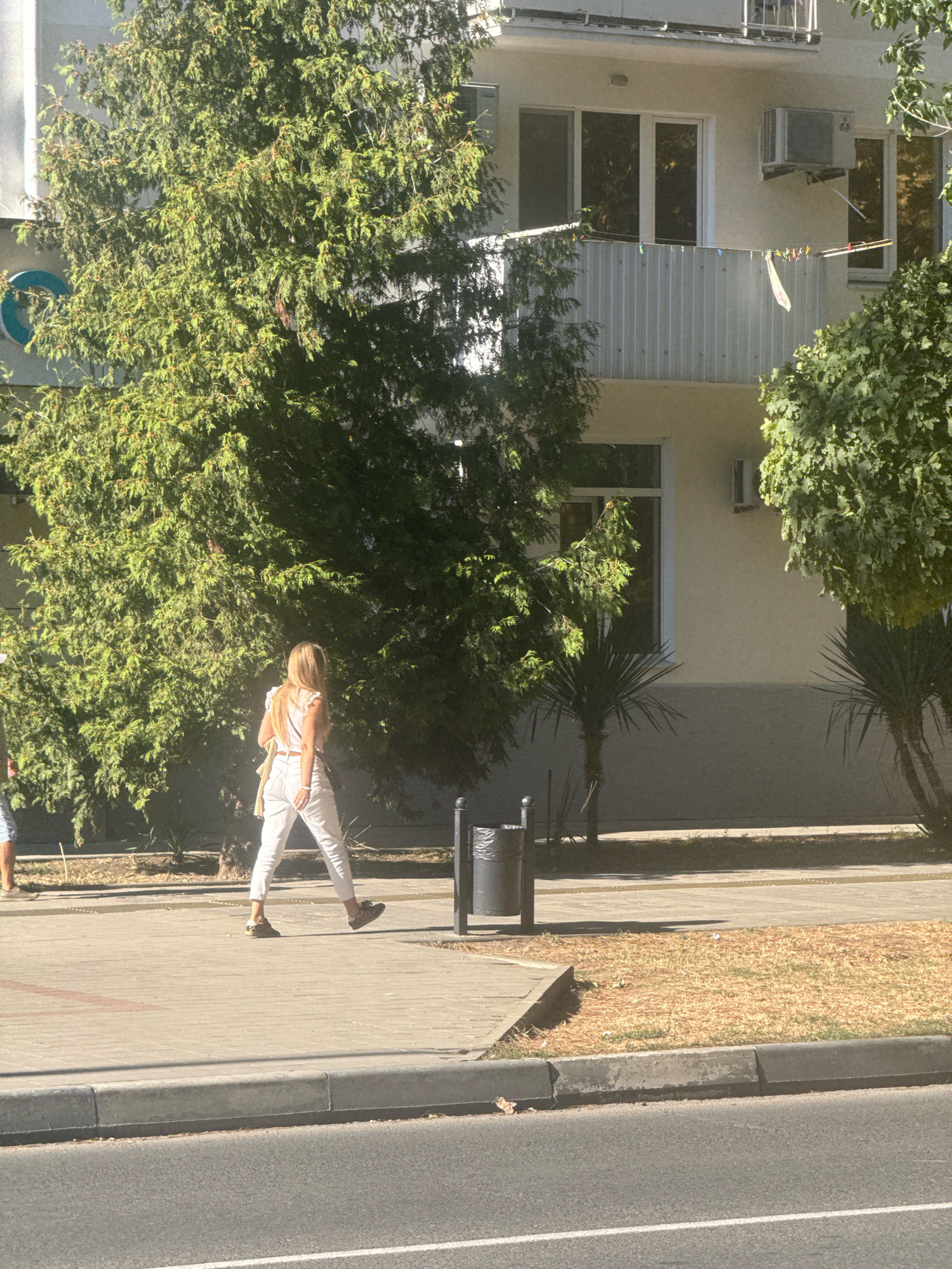 In Anapa, Russia, a woman with blonde hair, wearing a white top and white pants, walks along a sidewalk. She carries a tan bag over her shoulder. Behind her, a large green tree provides shade. In the background, a building with a balcony displays laundry hanging on a clothesline, and an air conditioning unit is visible. A trash can stands near the sidewalk. The scene is illuminated by bright sunlight.