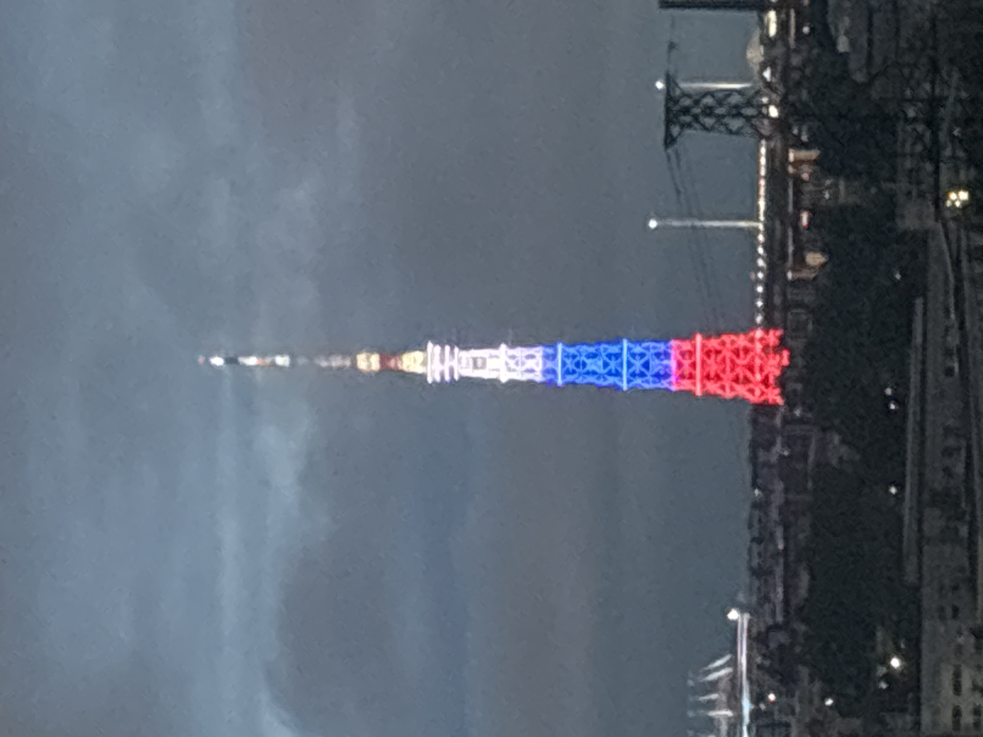 A distant view of a tall tower illuminated with red, white, and blue lights, possibly depicting the French flag, on the right side of the picture. Dark city structures and buildings are near the tower, and a cloudy sky is above. The scene is likely a nightscape in an urban setting.