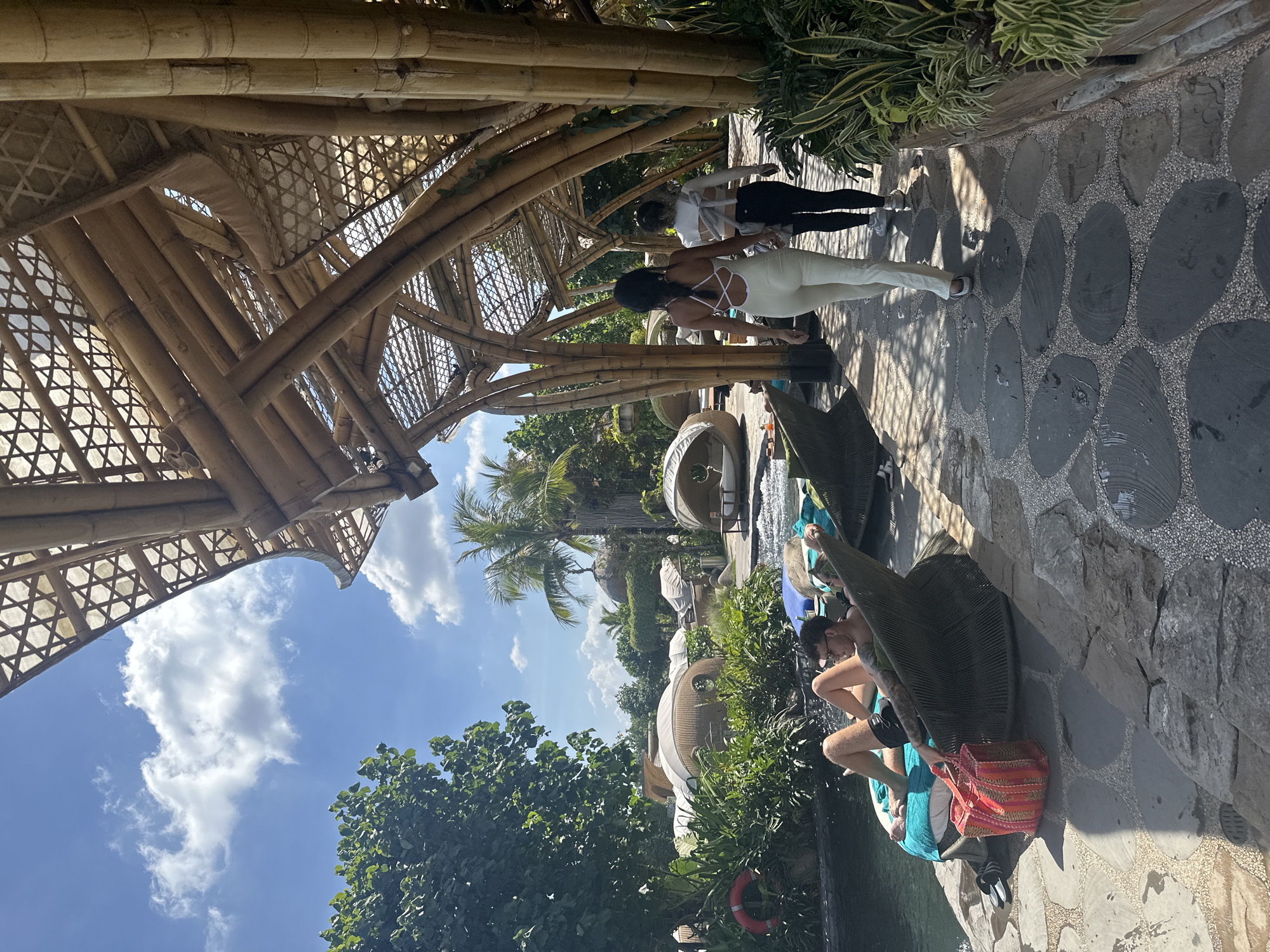 The image captures a sunny day at a resort in Kediri, Indonesia, characterized by a distinctive open-air pavilion constructed from bamboo. The pavilion's roof is intricately woven with bamboo, supported by large bamboo poles, creating a semi-transparent canopy that allows sunlight to filter through. A paved walkway, made of irregular dark stones interspersed with small pebbles, runs alongside the pavilion.
On the left side of the walkway, a person is relaxing on a woven lounge chair by a pool of water. The individual is wearing shorts and sunglasses, with tattoos visible on their arms. Next to the chair is a brightly colored striped tote bag. In the background, lush green foliage, palm trees, and uniquely shaped dome-like structures are visible, suggesting a tropical resort setting.
Two individuals are walking away from the camera on the pathway, heading towards the right side of the frame. The person in front is wearing a white sleeveless jumpsuit with a criss-cross back detail, while the person behind them is wearing a white long-sleeved top and black leggings.
The sky is a clear blue with scattered white clouds. The overall scene evokes a relaxed and tropical ambiance.