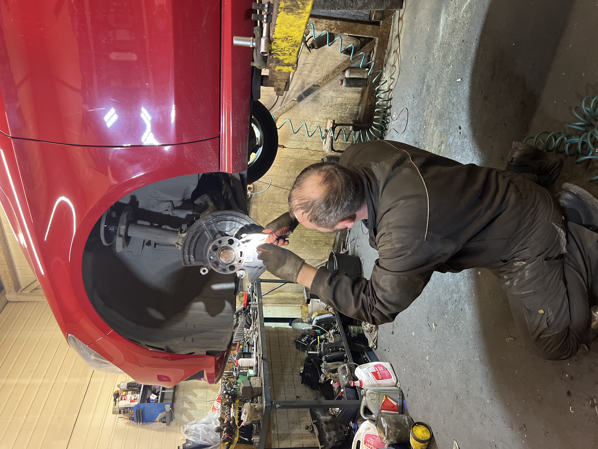 An automotive mechanic is captured working on the front wheel assembly of a red car inside a garage in Vsevolozhsk, Russia. The car is elevated on a yellow metal car lift, with its front right wheel removed, exposing the hub and brake assembly.
The mechanic, a middle-aged man with a balding head, is kneeling on the concrete floor, intently focused on the car's hub. He is wearing dark work overalls and protective gloves. His hands are positioned near the hub, suggesting he is inspecting, cleaning, or performing maintenance with a small tool or light, which reflects off the metal surface.
The garage environment is visible around him, featuring a cluttered but organized workspace. Shelves filled with various bottles and containers, including those labeled "COOLANT" and potentially "OIL," are seen behind the mechanic. Teal-colored coiled air hoses snake across the dirty concrete floor. The overall lighting suggests an indoor setting with artificial illumination. The red car's bodywork shows some reflections from overhead lights and minor dust or scuffs. The floor appears worn and possibly stained with grease or oil, typical of a workshop.
