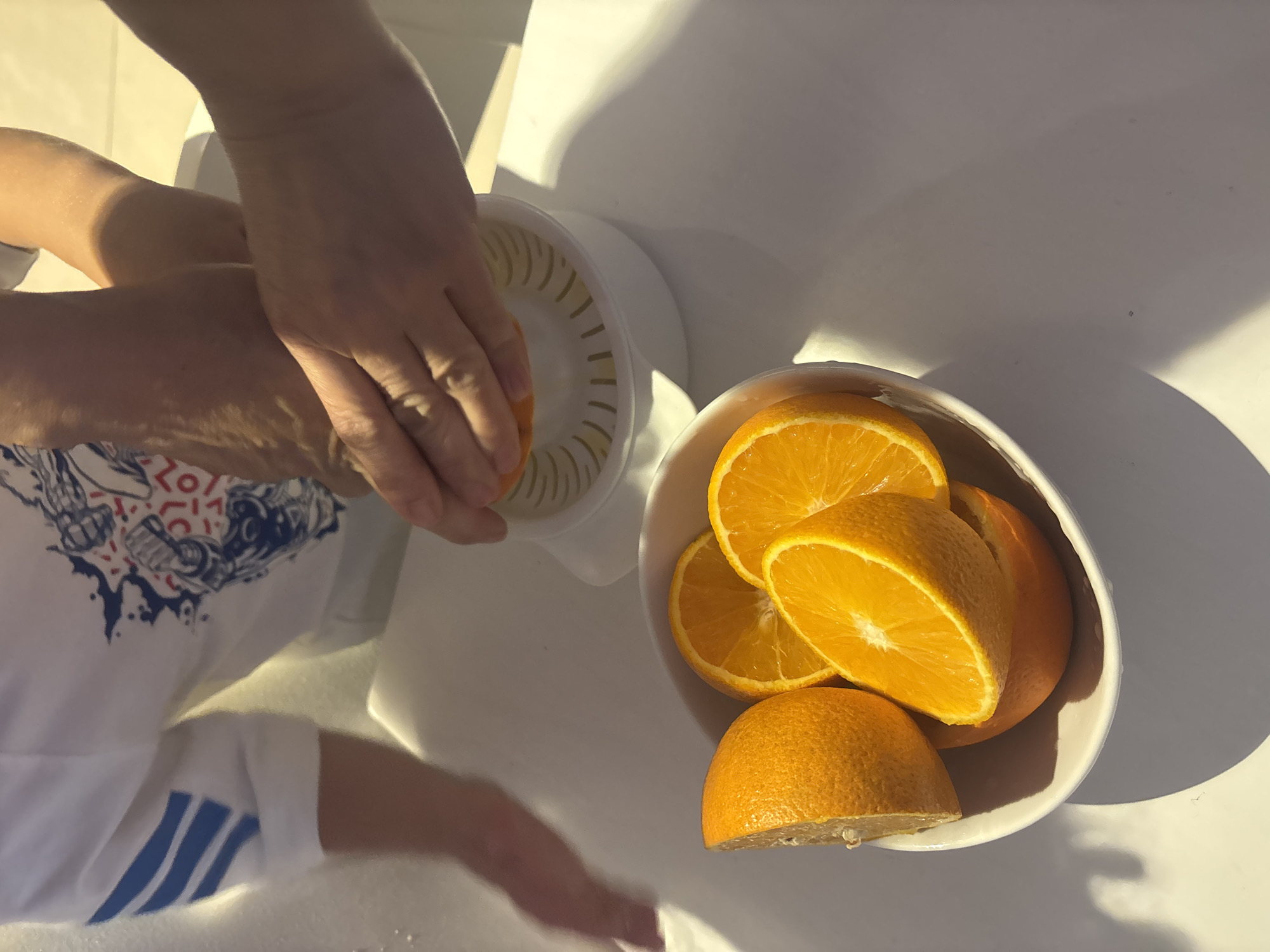 The image depicts a close-up, overhead view of hands using a manual citrus juicer to extract juice from an orange. Several halved oranges are visible in a white bowl in the foreground. The background shows parts of a person wearing a white t-shirt with blue stripes and a graphic design. The scene appears to be indoors, possibly in a kitchen, with bright lighting creating shadows on the white surface. There is no visible indication of the location being in Dubai, United Arab Emirates.