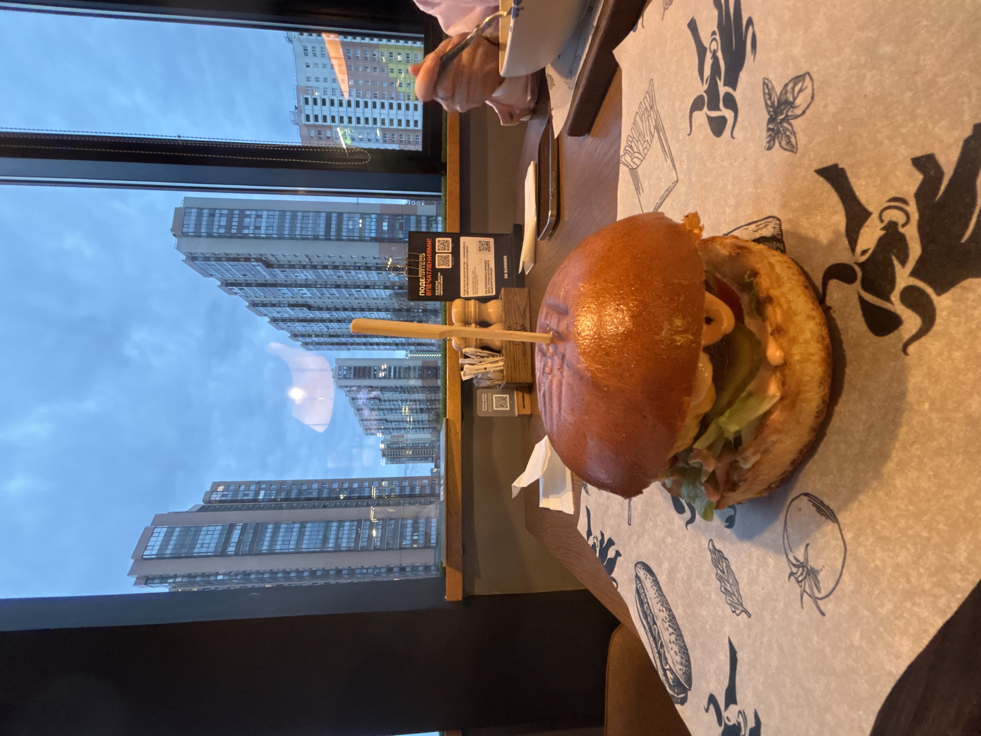 The image shows a burger on a table with a scenic view from a window. Outside the window, high-rise apartment buildings dominate the view under a cloudy sky. The setting is urban, likely in a densely populated area.
The burger appears to be freshly prepared, held together by a wooden stick. The table has a decorative paper liner with illustrations of food items and figures. The location is Saint Petersburg, Russia, indicated by the "SB Burgers" signage and the "ПОДЕЛИТЕСЬ ВПЕЧАТЛЕНИЯМИ!" text.
