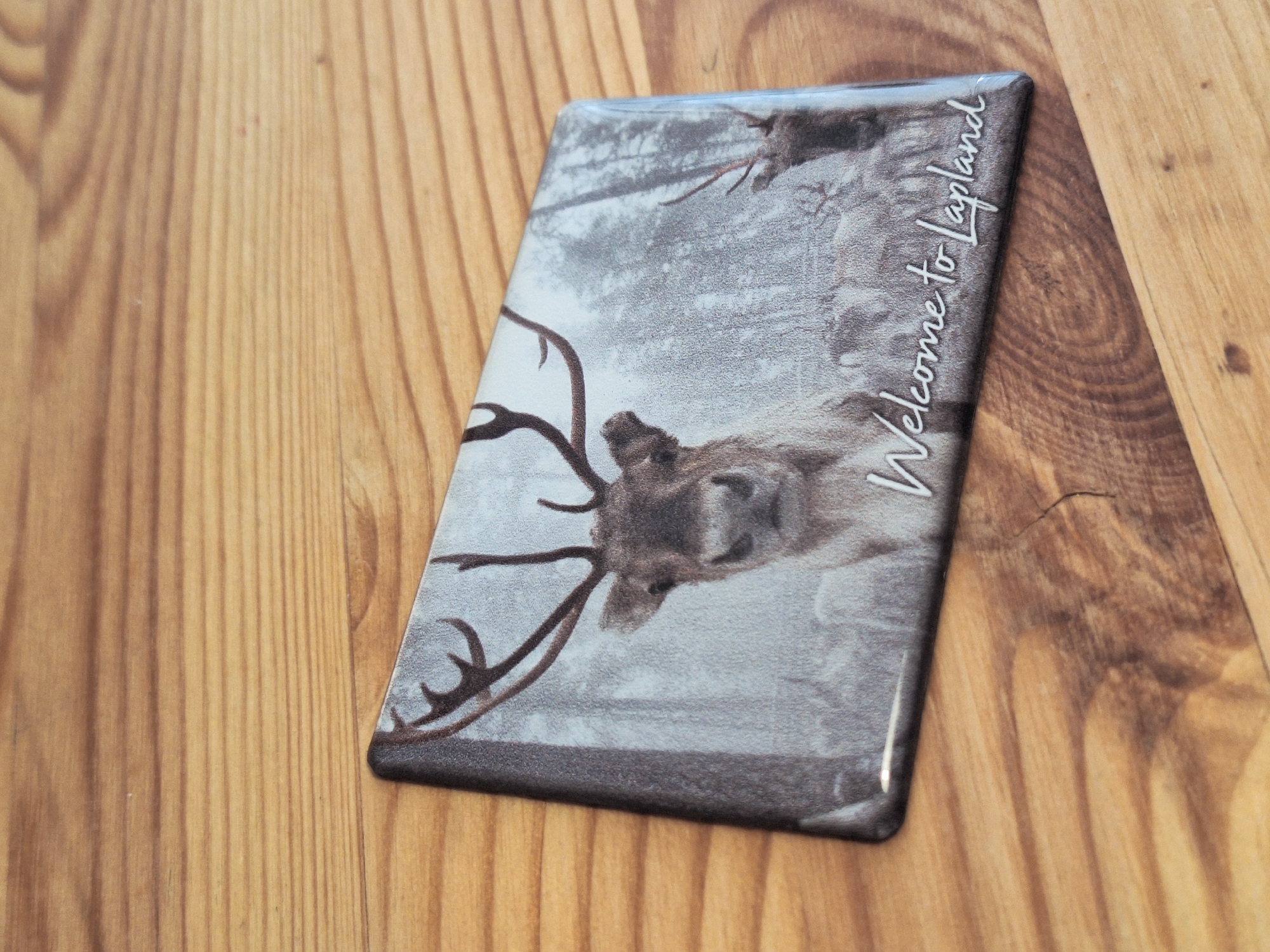 A rectangular object, likely a souvenir magnet or coaster, is prominently featured in the image, positioned diagonally on a wooden surface. The object has a glossy finish, reflecting light, and appears to be made of or coated with resin.
The surface beneath the object is a light to medium-toned wooden board or table, displaying clear, linear wood grain patterns that run vertically and diagonally, indicating natural wood planks. Some minor imperfections or faint marks are visible on the wood.
The object itself showcases a desaturated or grayscale image of a reindeer, or a similar deer species, with large, prominent antlers. The reindeer's head and upper body are visible, looking directly at the viewer with a somewhat soft expression. The background of the image suggests a sparse, wintery forest or a snowy landscape with indistinct trees.
On the right side of the object, in a flowing, white cursive font, the text "Welcome to Lapland" is clearly visible. This text and the reindeer imagery strongly suggest a theme related to northern, cold regions.
No specific activity is taking place; the image is a static shot of a decorative item. The overall impression is that of a souvenir or keepsake from a northern destination. While the object explicitly references "Lapland," often associated with Scandinavia, it aligns with the general theme of cold-climate nature found in regions like Chaikovsky, Russia, where such northern-themed souvenirs would be common.