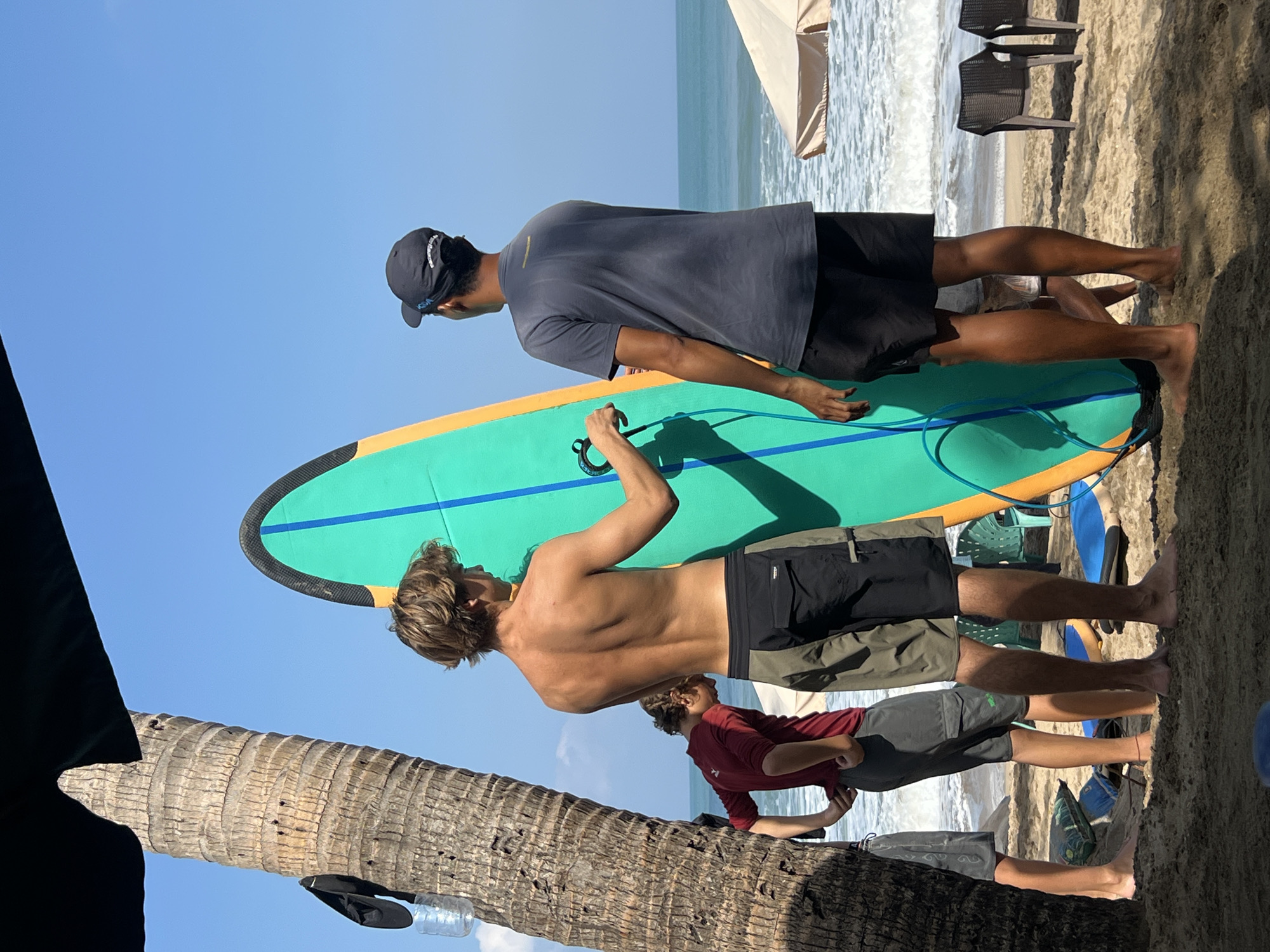 At the beach in Kuta, Indonesia, four males are present near the shoreline. A tall palm tree with rough bark stands to the left of the frame, partially shaded by a dark awning. A large, bright green surfboard with orange trim and blue lines is held upright by a shirtless male with blonde hair, who is wearing black and grey shorts. He is facing away from the camera, holding the surfboard with his right hand. To his right, another male wearing a dark grey t-shirt, black shorts, and a black baseball cap is also facing away, looking towards the ocean. A third male, wearing a red t-shirt and grey shorts, is positioned between the palm tree and the surfboard, also facing away, with his head down. A fourth male is partially visible on the far left, wearing grey shorts. The ocean waves are visible in the background, with white surf breaking. Two beach chairs and a beige umbrella are also present on the sand further down the beach. The sand is light brown and has shadows cast on it. The sky is a clear blue.