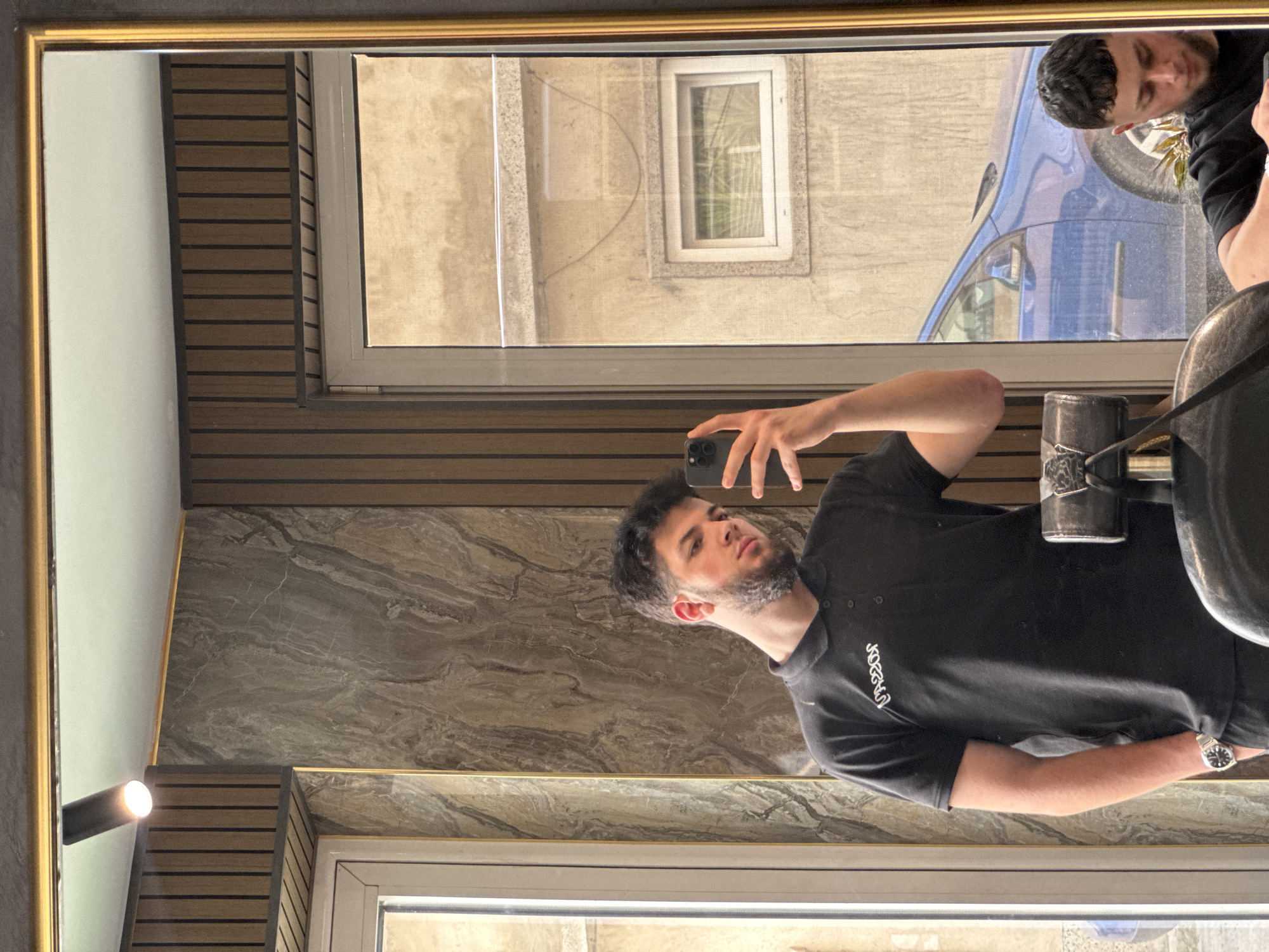 The image, rotated 90 degrees clockwise, captures a man taking a mirror selfie in an indoor setting in Varna, Bulgaria. The main subject is a young man with dark hair and a beard, wearing a black polo shirt with stylized white text, possibly "Unicorn," embroidered on the left chest. He has a metallic watch with a round face on his left wrist. He is holding a dark-colored smartphone in his right hand, positioned to capture his reflection.
The mirror has a gold-colored frame and reflects a contemporary interior. Visible elements within the reflection include a marble-patterned wall, brown wooden slatted paneling, and a wall-mounted light fixture emitting a bright glow. Through a white-framed window also reflected in the mirror, an outdoor scene is visible, showing parts of a building with light-colored concrete facades and a blue car partially visible, suggesting a street or parking area. The lighting indicates it is daytime with clear weather. The man maintains a neutral expression while taking the photo. The overall scene suggests a modern and well-lit interior space, possibly a lobby or a commercial establishment.