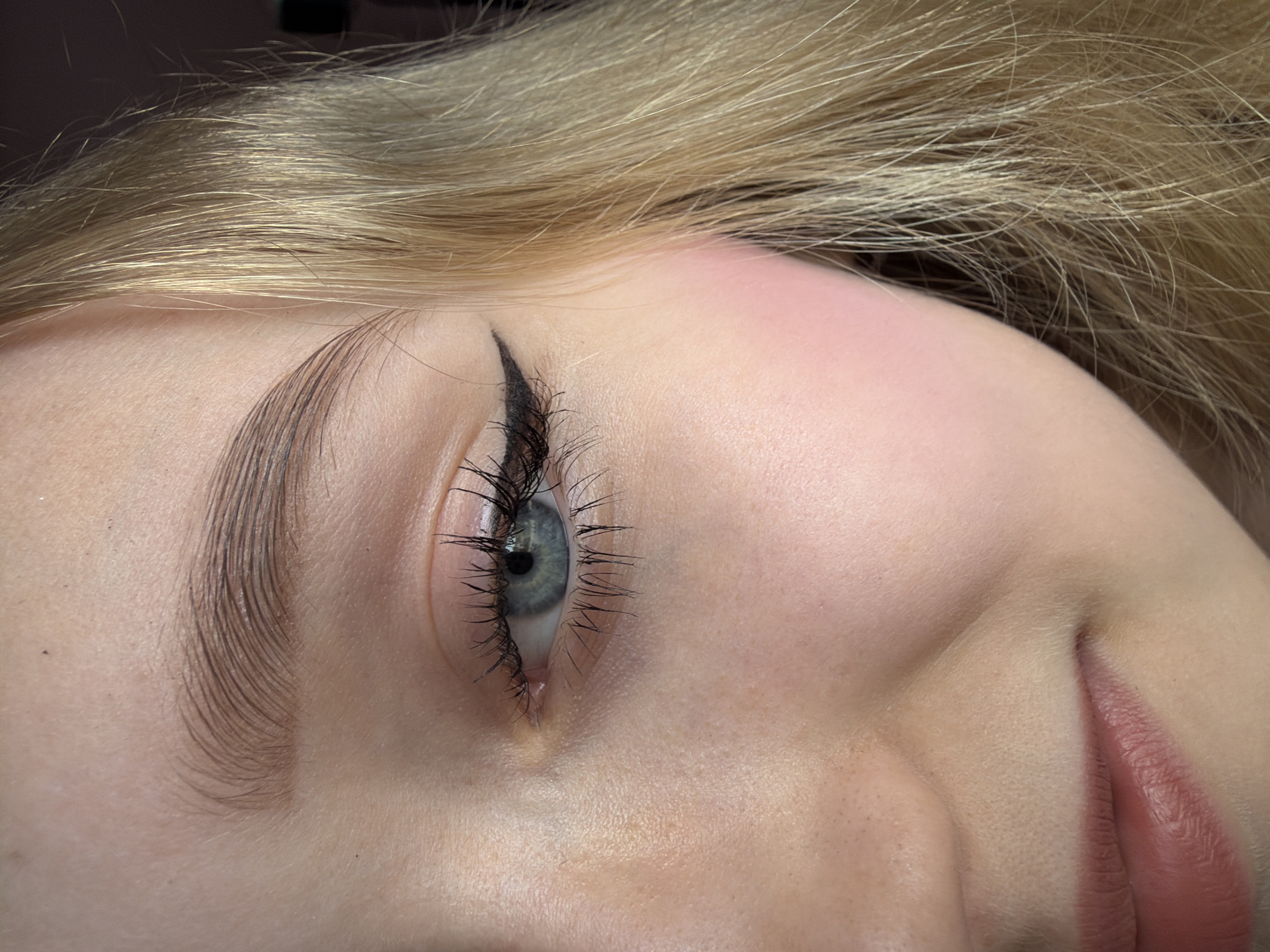 Close-up, angled view of a person's face. The person has blonde hair and fair skin. Their eye is open, with visible blue/gray iris, dark eyelashes, and eyeliner. The eyebrow is defined and groomed. Part of the nose and mouth are visible. The scene suggests a beauty or makeup setting but does not provide enough information to infer a specific location within Dreiliņi, Latvia.