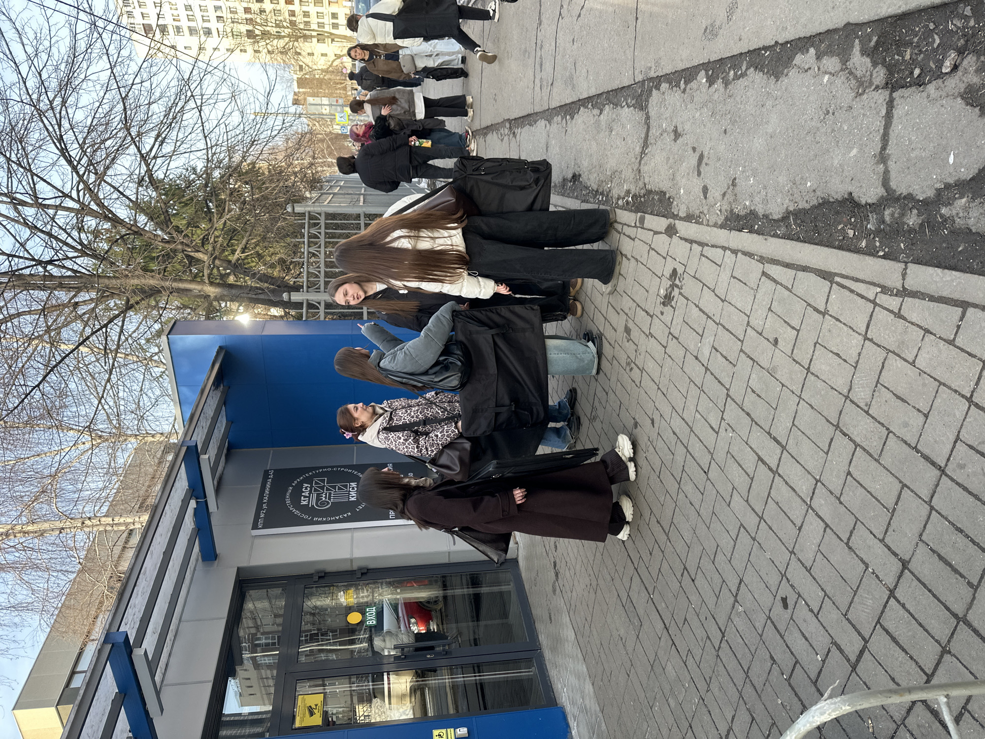 The image depicts an outdoor urban scene on a clear day at 43 Kalinin Street, Kazan, Russia, specifically outside Entrance 2 (КПП №2) of the Kazan State University of Architecture and Civil Engineering (КГАСУ/КИСИ). The setting is a paved sidewalk adjacent to a modern building with blue and grey architectural elements.
Six young females are grouped on the grey block-paved sidewalk near the building's glass double-door entrance.
1. A female on the far left wears a dark brown long coat, dark pants, and white sneakers, carrying a dark shoulder bag. She faces right.
2. Next to her, a female in a light grey puffer jacket and light blue jeans gestures with her right hand while holding a large, black, soft-sided case. She faces the group.
3. A third female, with long dark hair, wears a black jacket over a white hoodie and dark pants.
4. Centrally positioned, a female with long light brown hair wears a white puffer jacket, black wide-leg pants, and carries a large black soft-sided case on her left side, accompanied by a brown shoulder bag. She faces left, toward the other women.
5. To her right, a female with light brown hair and a pink hair clip wears a leopard print puffer jacket and light blue jeans, also holding a large black soft-sided case. She faces right.
6. The sixth female, partially visible from the back, wears a light grey jacket and blue jeans.
The females appear to be conversing; several are equipped with large, black, flat cases, likely for architectural drawings, art portfolios, or musical instruments.
The building features a multi-pane glass entrance door marked with "ВХОД" (Entrance) and a security camera decal. An overhead awning with blue and grey structural elements extends from the building. A sign on the building clearly identifies "КПП №2, ул. КАЛИНИНА Д.43" and the university name in Cyrillic.
Beyond this group, the sidewalk continues to a rougher asphalt strip and a wider asphalt area. Numerous other pedestrians are visible in the mid-ground and background, walking along the urban thoroughfare, indicating a moderately busy street. Bare deciduous trees and some evergreen foliage line a metal fence separating the sidewalk from a greener area. Tall multi-story buildings are present in the distant background. A "P" (Parking) sign is partially visible. The overall scene suggests a weekday, possibly late morning or afternoon, given the pedestrian activity.
