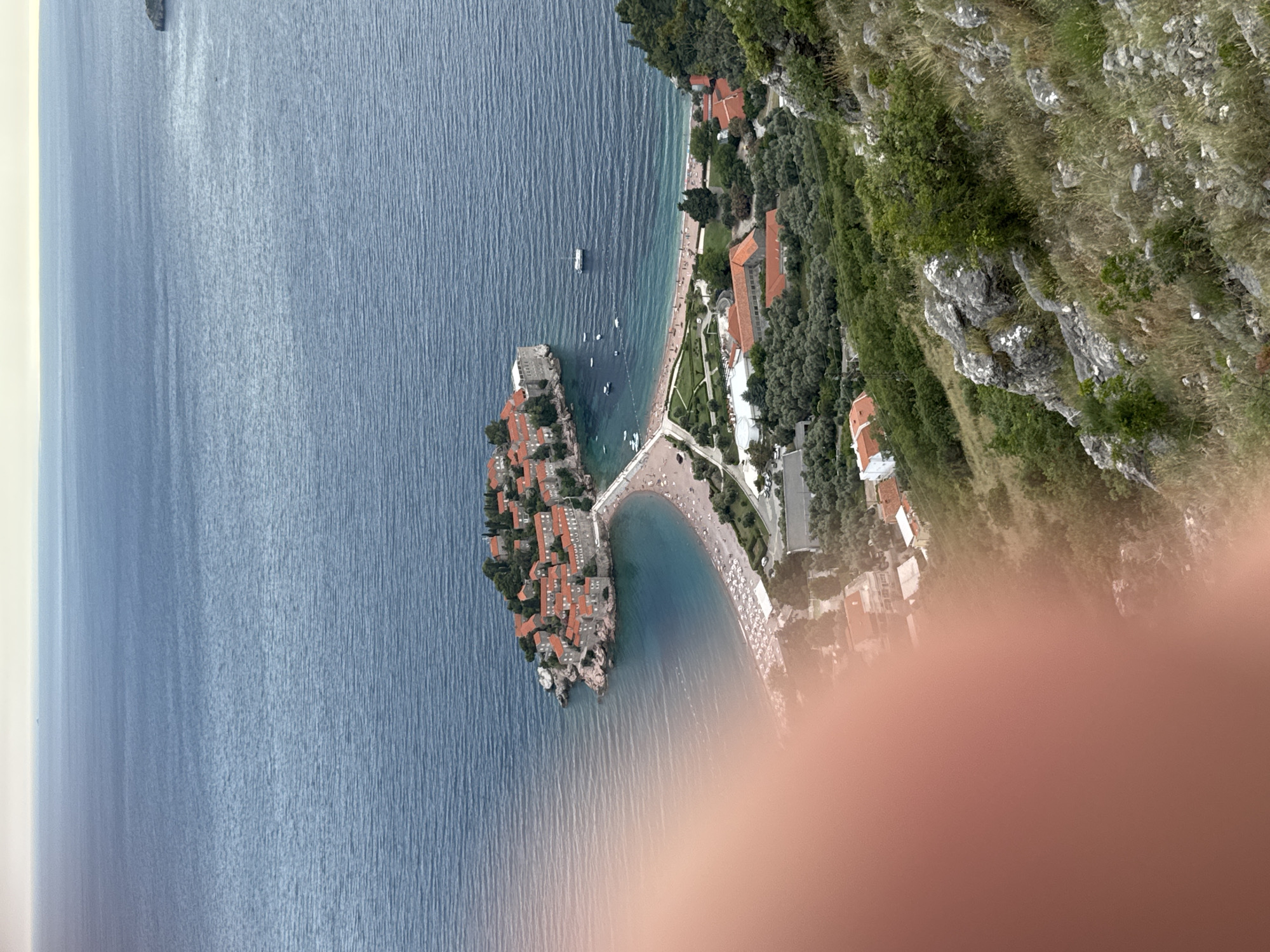 This aerial photograph depicts the picturesque coastal town of Sveti Stefan, Montenegro. The primary subject is the iconic island of Sveti Stefan, a fortified village connected to the mainland by a narrow causeway. The island is densely packed with terracotta-roofed buildings, nestled amongst lush green trees.
The scene is set against a backdrop of the clear, blue Adriatic Sea, which is calm with subtle ripples on its surface. Several small boats are visible dotting the water near the island and along the coastline. The mainland features a sandy beach dotted with numerous white umbrellas, suggesting a warm, sunny day, likely in the late morning or early afternoon given the lighting. Adjacent to the beach are hotels and resort buildings with similar terracotta roofs, surrounded by dense greenery and winding roads.
The photograph captures the natural beauty of the Montenegrin coast, with the rugged, grassy hills rising behind the developed area. The overall impression is one of a tranquil and idyllic vacation destination. A large, blurred area of skin tone in the foreground suggests the photographer's hand or finger may have been in front of the lens at the moment the photo was taken, partially obscuring the lower right portion of the scene. There is no visible text in the image.