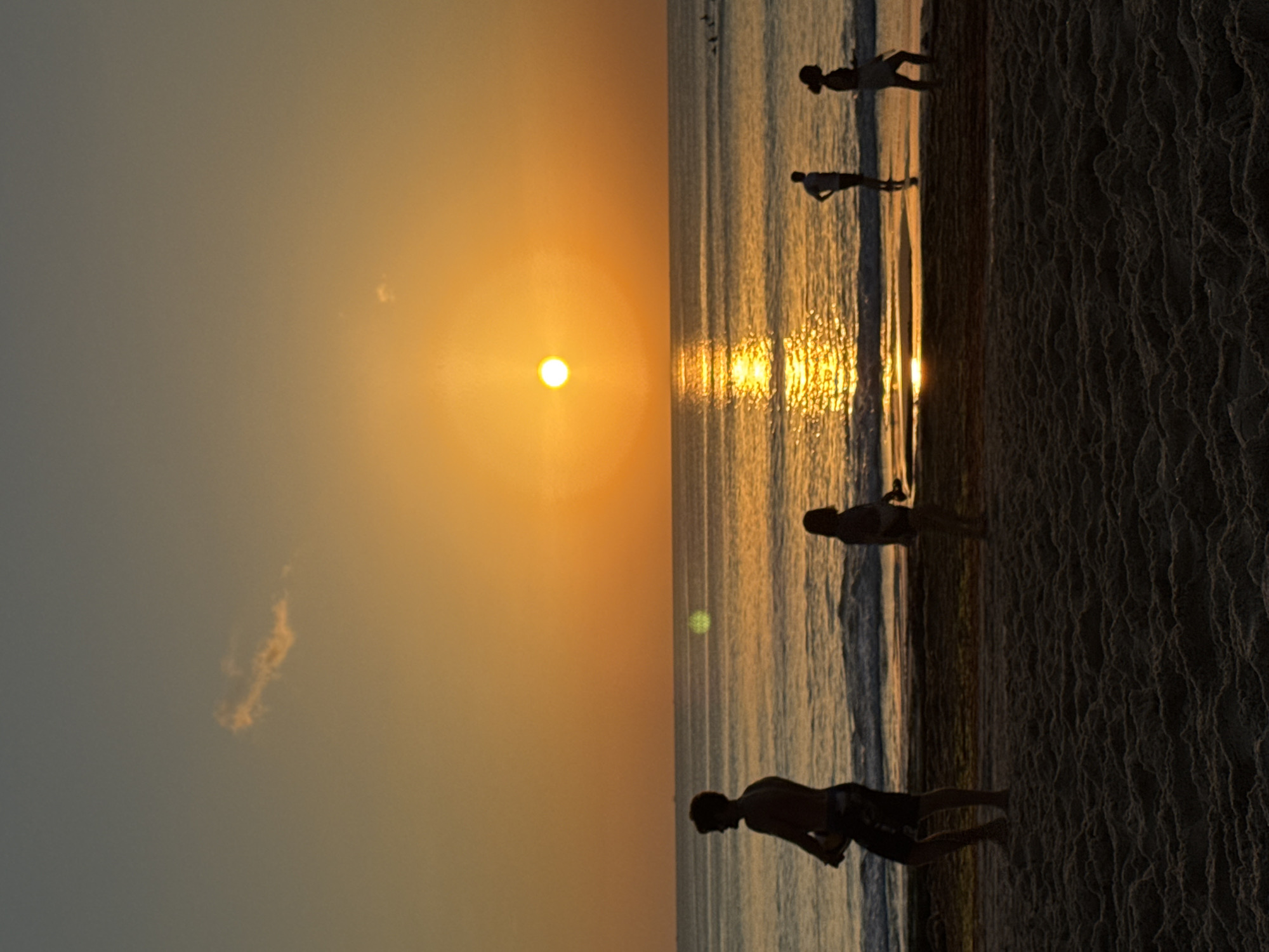 This image depicts a beach scene at sunset in Kuta Selatan, Indonesia. Silhouetted figures are present on the sand and in the shallow water. In the foreground, a man stands facing left, holding a circular object, possibly a frisbee or a ball. To his right, a woman stands facing the ocean, with her hands at her sides. Further out in the water, two more silhouetted figures are visible. The sun is setting over the horizon, casting a warm orange glow across the sky and reflecting on the calm ocean waves. A single wispy cloud is visible in the sky. The beach is covered in dark sand with a textured appearance.