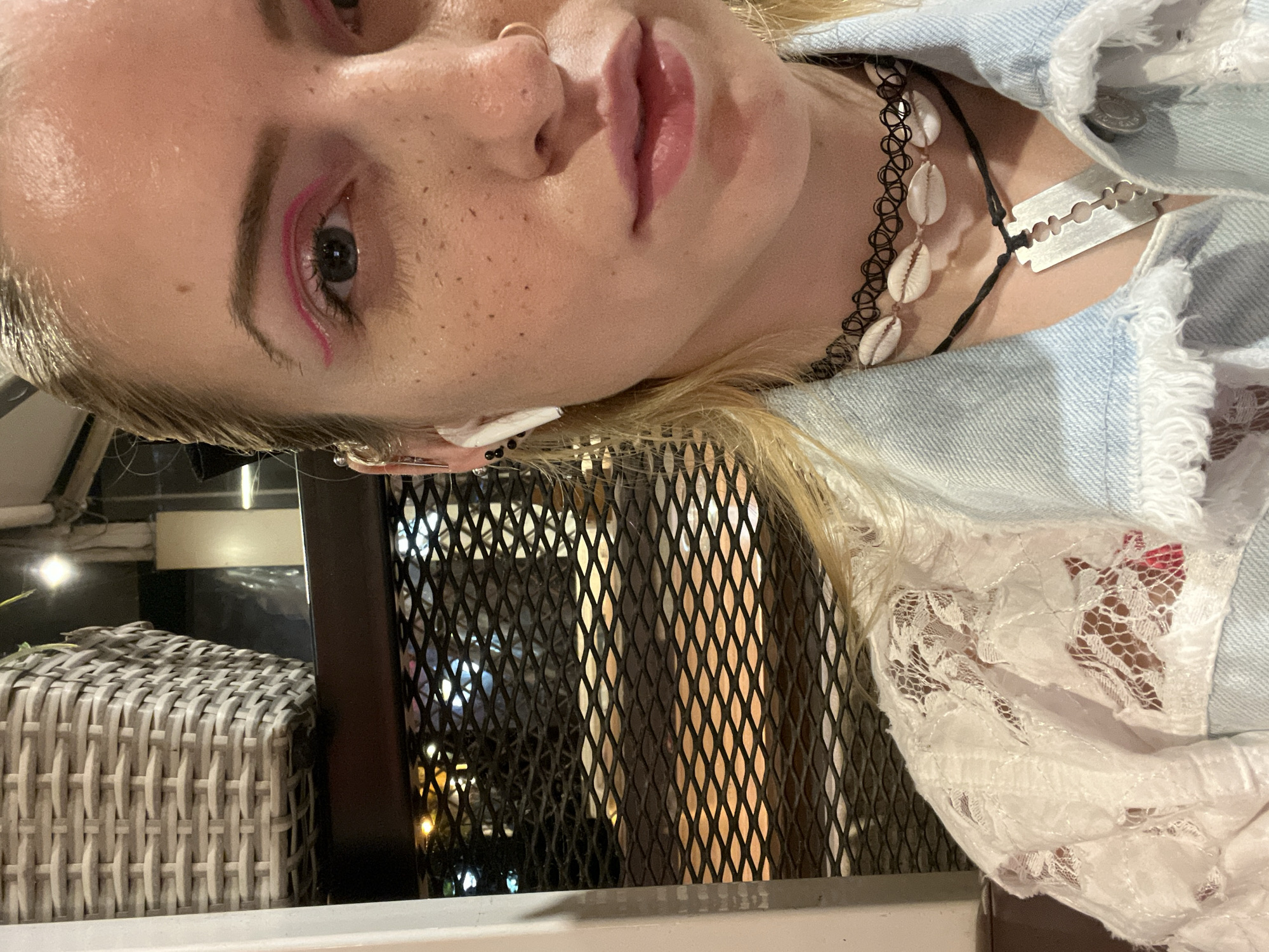 A close-up, slightly angled image shows a person with light skin and blonde hair, adorned with freckles, a nose ring, and striking pink eyeliner. They are wearing a blue denim jacket over a white lace top. Around their neck are two necklaces: one a black elastic choker with white shells, and the other a thin black cord with a silver razor blade pendant.
Behind the person, there is a wrought iron-style screen or barrier, and to the left, a woven grey and white container, likely a planter or basket. The background suggests an outdoor setting, possibly a patio or restaurant area.
While specific location cannot be determined, the question refers to city of Plovdiv, Bulgaria.