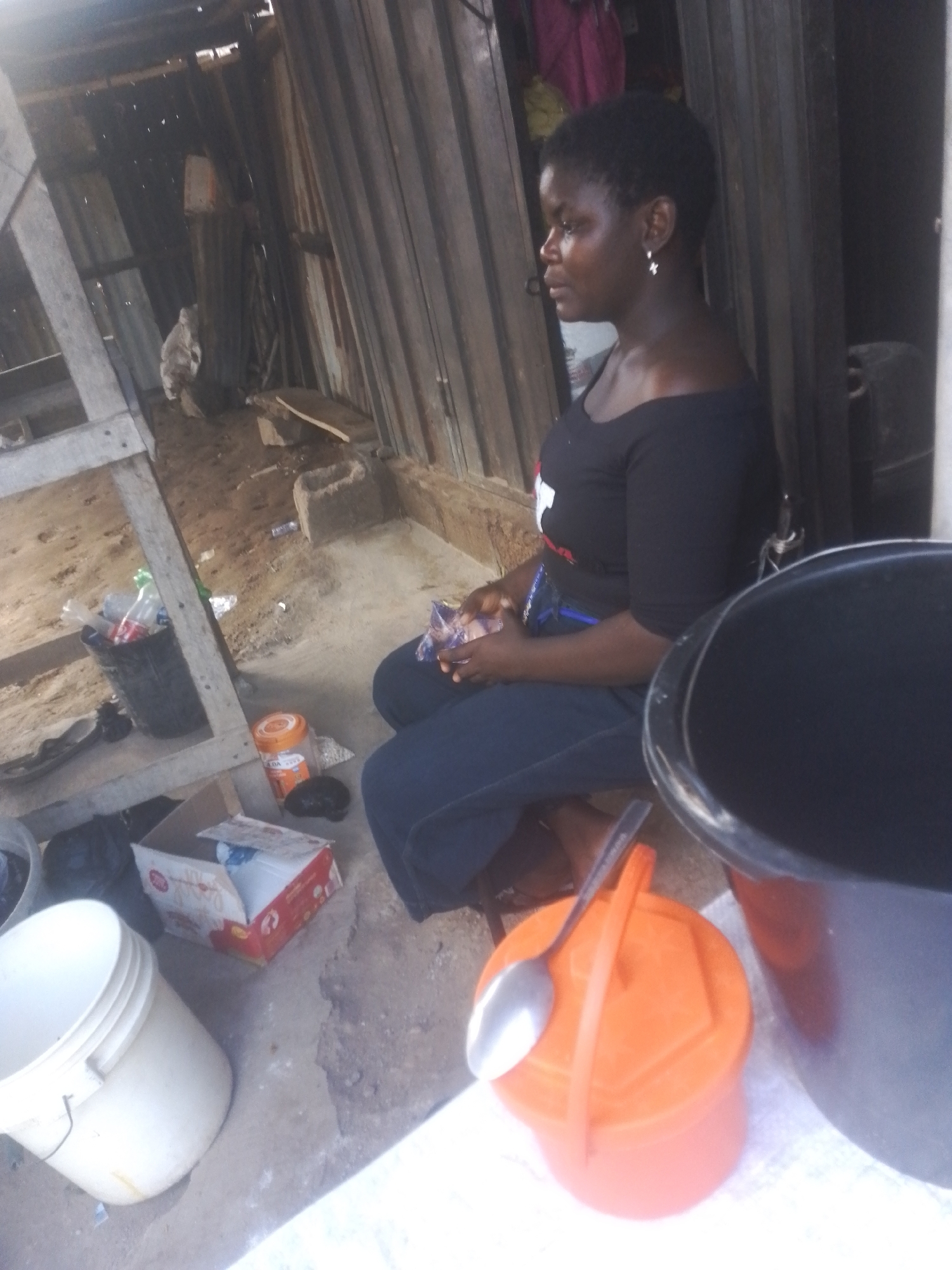 A medium shot captures an African woman sitting in an outdoor, rustic setting. She is wearing a black off-the-shoulder top and dark blue pants, with a small silver earring in her visible ear. Her short, dark hair frames her profile as she looks to her left. The woman holds a small plastic bag in her hands.
The environment appears to be a makeshift shelter with corrugated metal walls and wooden structural elements. The ground is rough dirt and concrete. Various objects are scattered around her, including a white bucket, an orange plastic container with a spoon resting on its lid, and a red and white cardboard box. In the background, a black bucket and other debris are visible. The lighting suggests an overcast day.